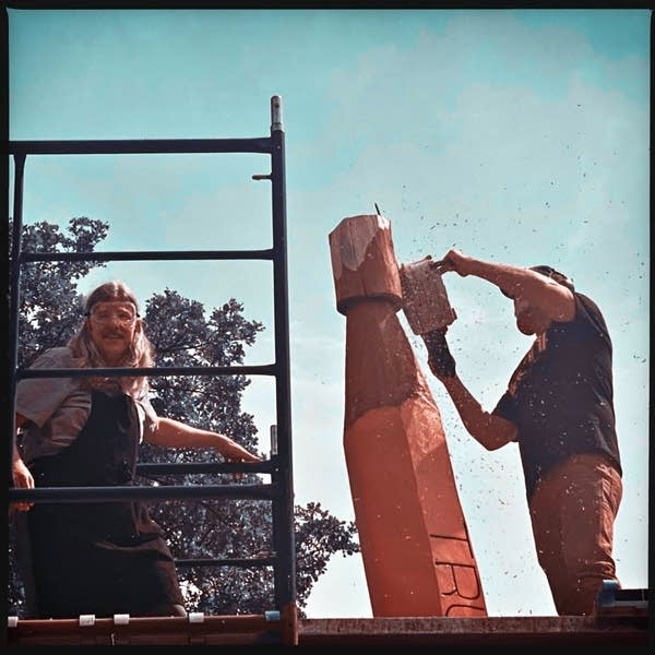 Person shaves wood sculpture, another stands on ladder