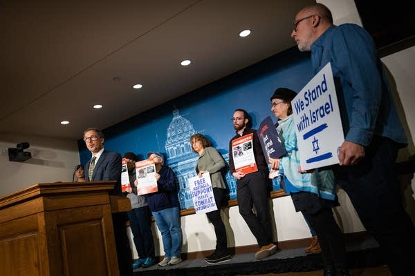 A man speaks at a podium as people hold signs behind him