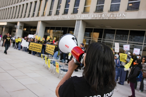 In this photo, demonstrators holding signs are standing outside the U.S. Department of Education's building in Washington, D.C., protesting against planned mass layoffs at the agency. In the foreground, a person is speaking into a megaphone.