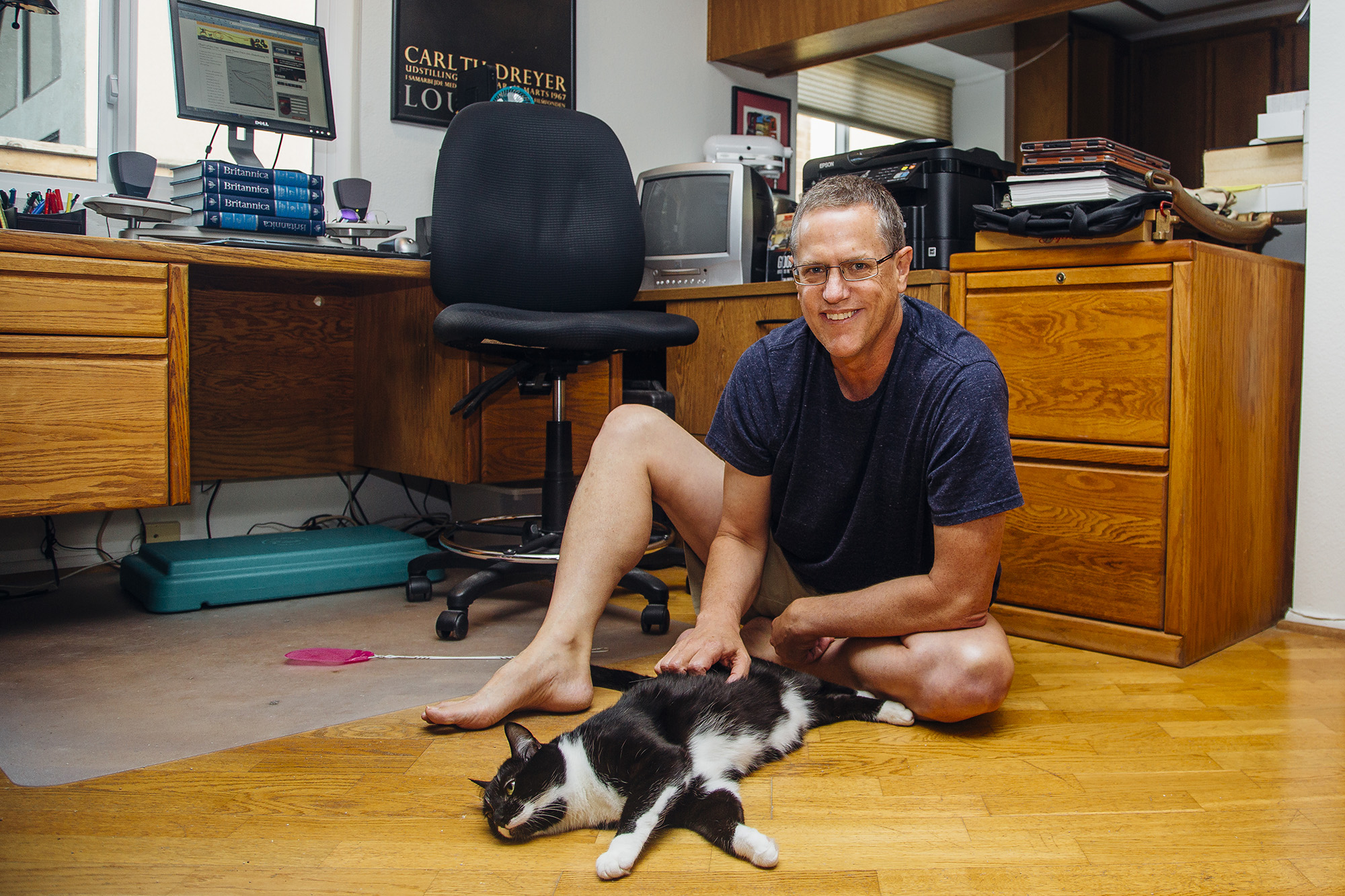 Smiling man sitting on floor of a home office, petting a black and white cat.