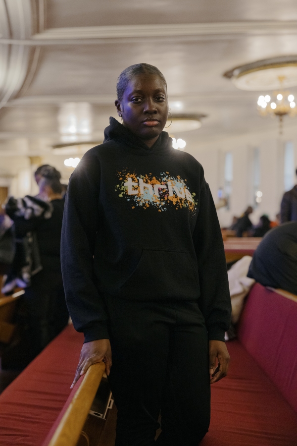 Janelle Davis, 16, stands near a church pew. She's wearing a sweatshirt that says, "Thrive."