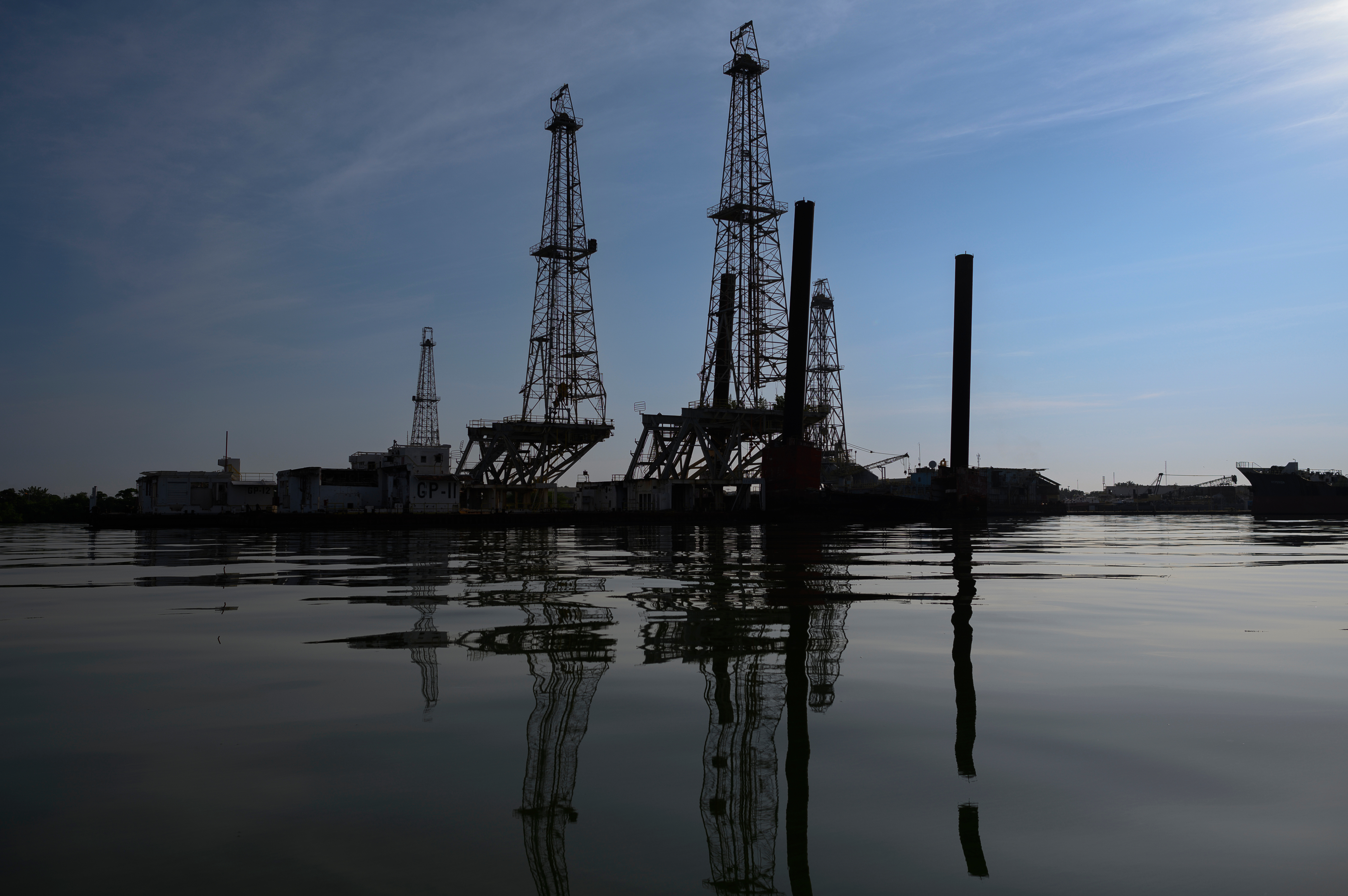 Oil derricks stand in sharp silhouette against a darkening sky, reflected in a large body of water