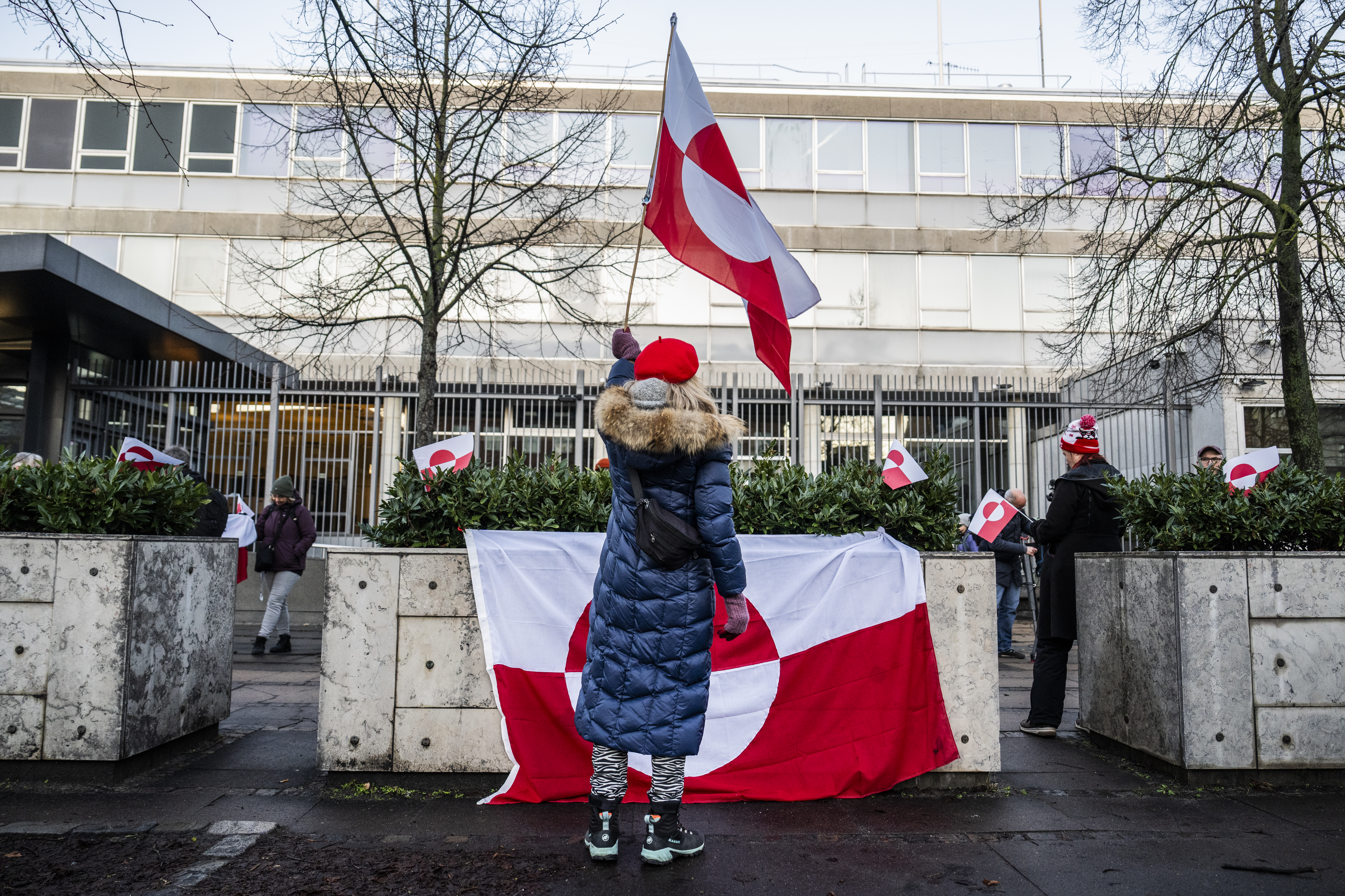 A woman holding the flag of Grenland and wearing a long blue coat with fur trim stands in front of a large Greenland flag hanging on concrete barriers 