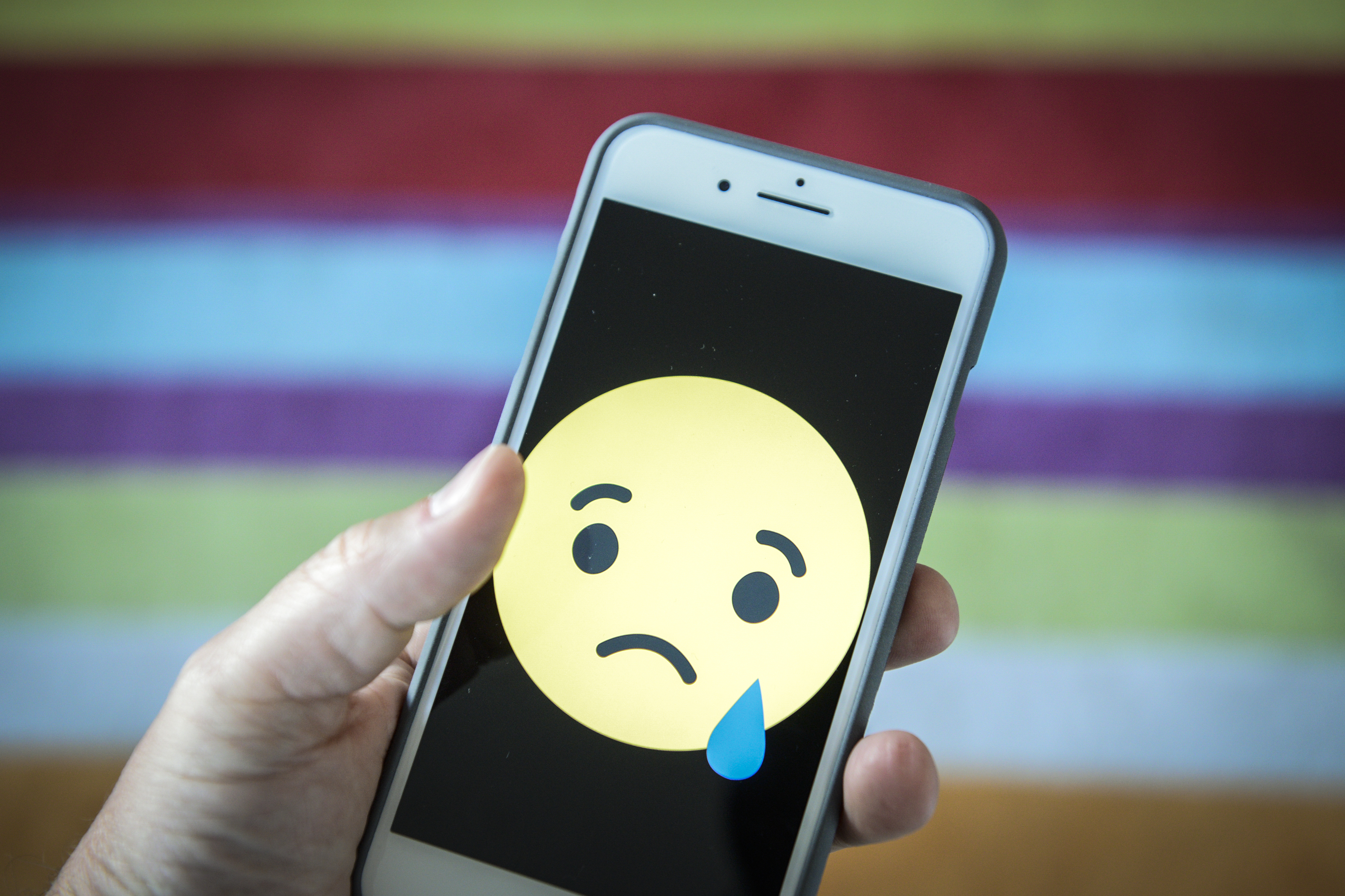 A hand holds a smartphone showing a large sad face emoticon against a black background.