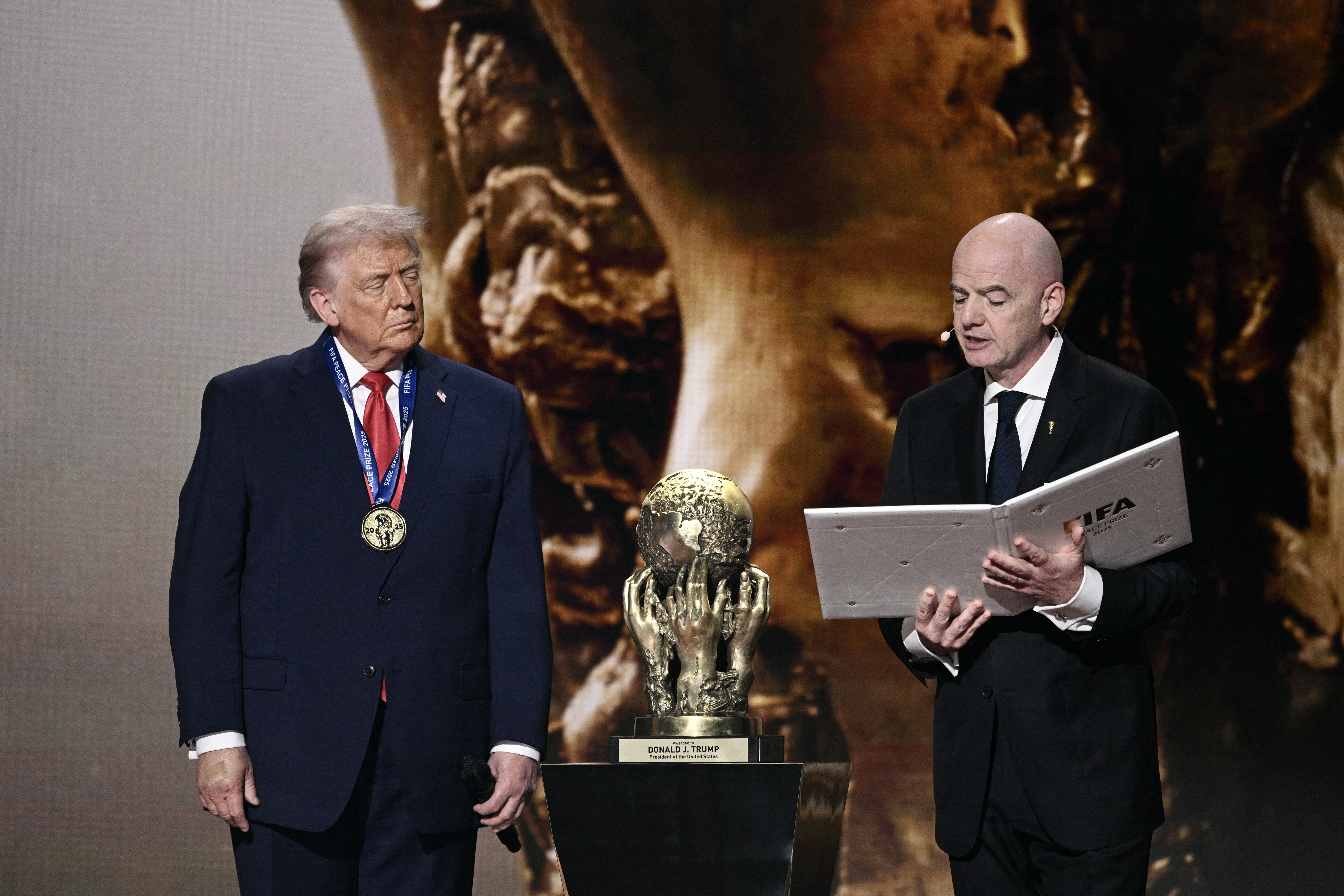 Trump, wearing a medal, stands next to FIFA President Gianni Infantino. Both men are wearing suits and there is a trophy between them