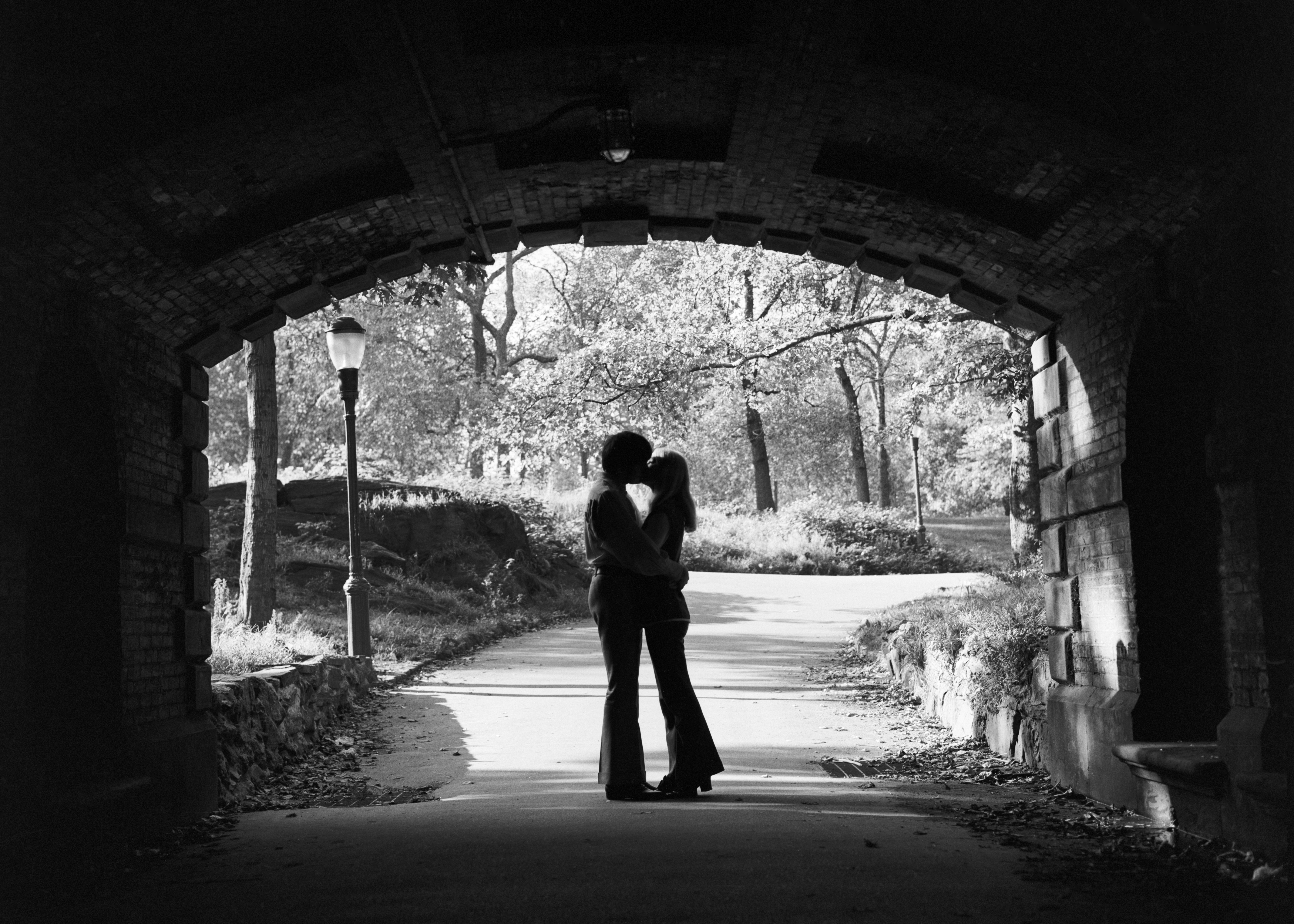 a black and white photo shows a silhouette of two anonymous people, one seems to be male-presenting and the other female-presenting, embracing within a public-walkway tunnel in Central Park, NYC 
