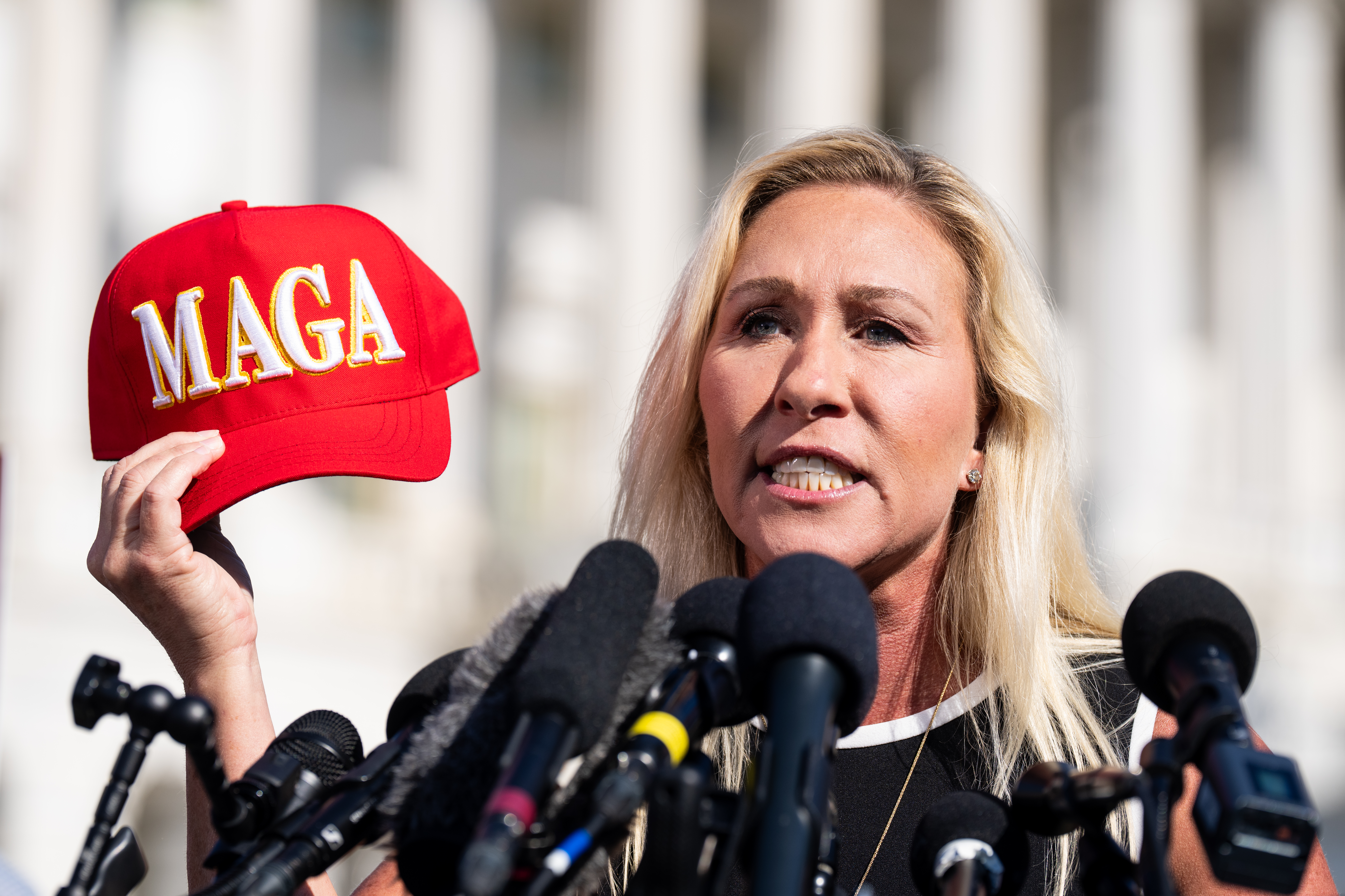 Rep. Marjorie Taylor Greene holds a red "MAGA" hat in front of an array of microphones, in front of the US Capitol Building.