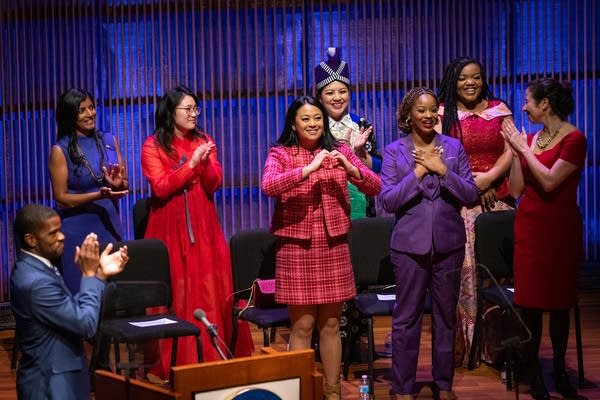 Seven women celebrate on a stage as a man claps