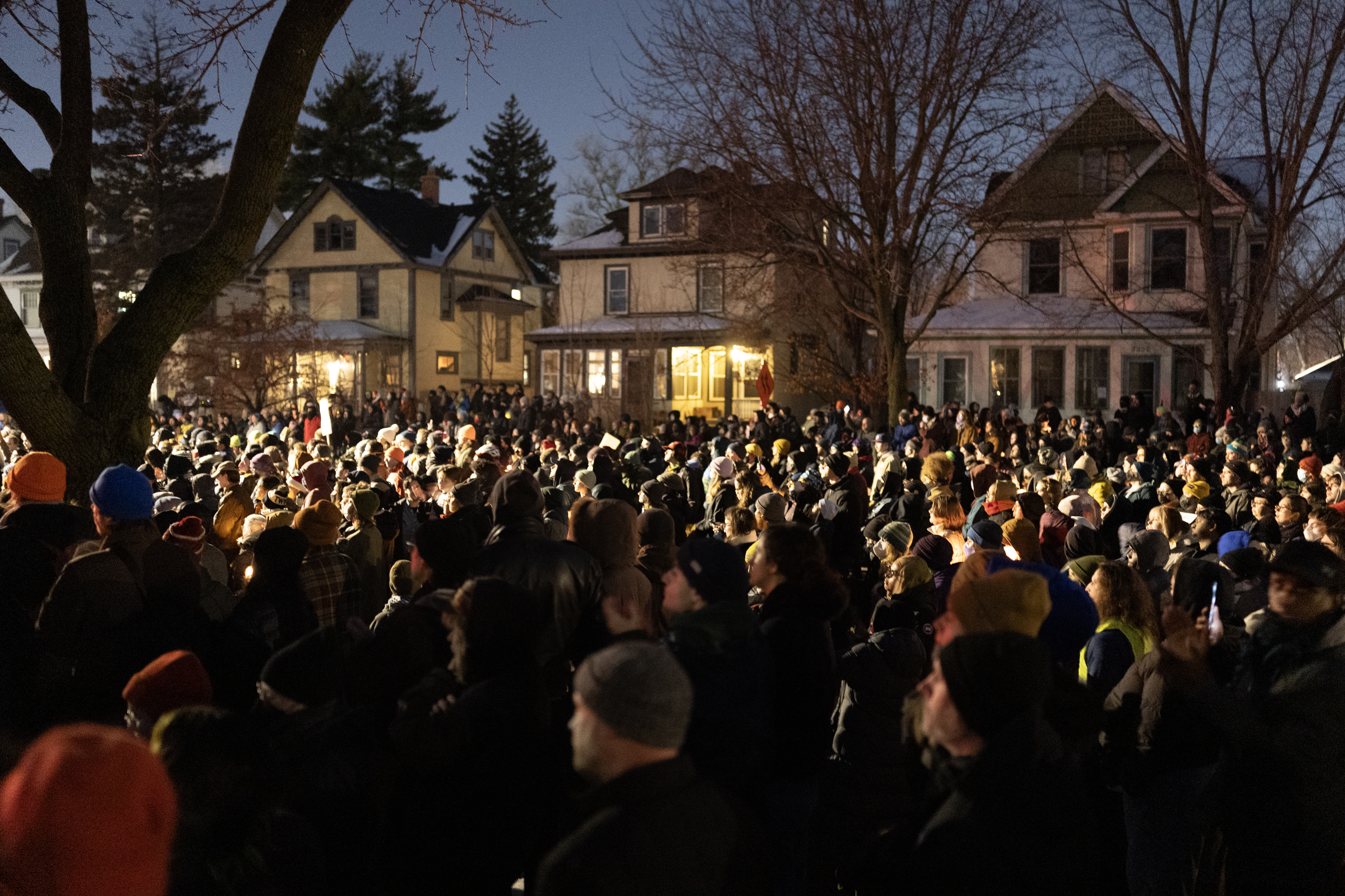 A large vigil at night, with many people in the crowd holding candles in the street with houses and trees in the background