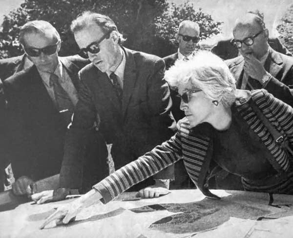 A black and white photo with a woman pointing at drawings on a table, men in suits surround her looking at the table.