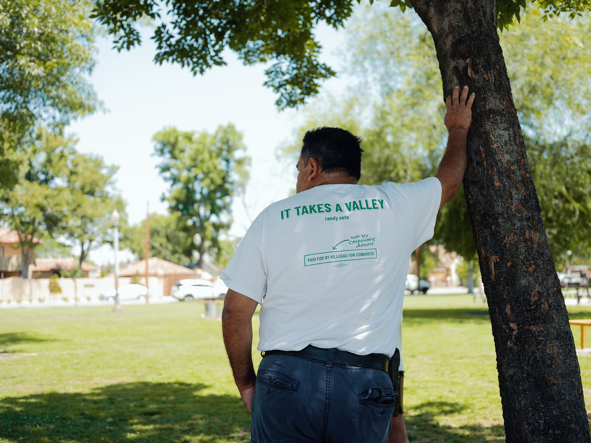 A man, seen from behind, leans against a tree in a park. The back of his T-shirt reads: "It takes a valley / randy.vote / Paid for by Villegas for Congress / Not by corporate donors."