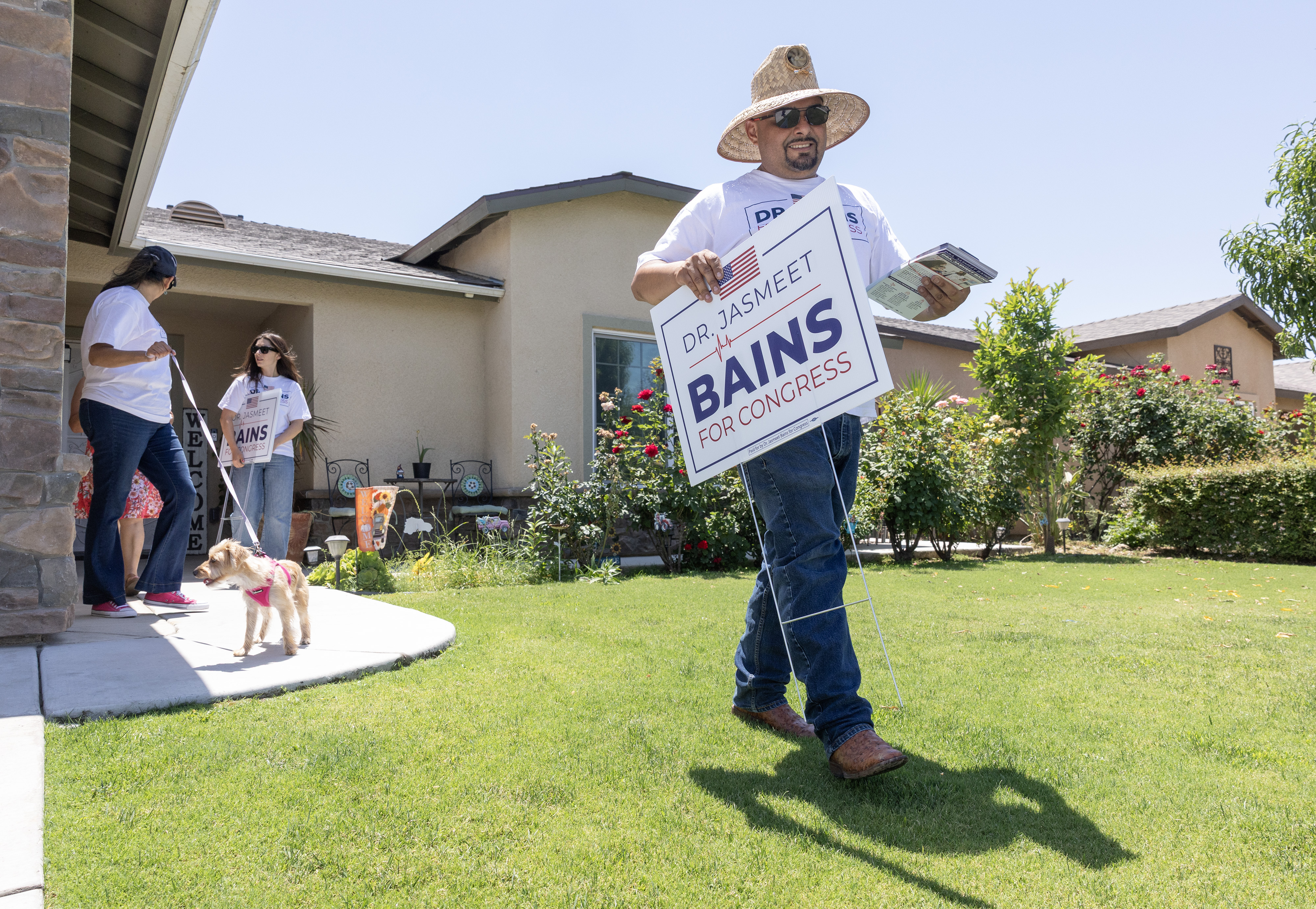 A man wearing a straw hat puts a walks across a home's lawn, holding a yard sign that reads, "Dr. Jasmeet Bains for Congress."