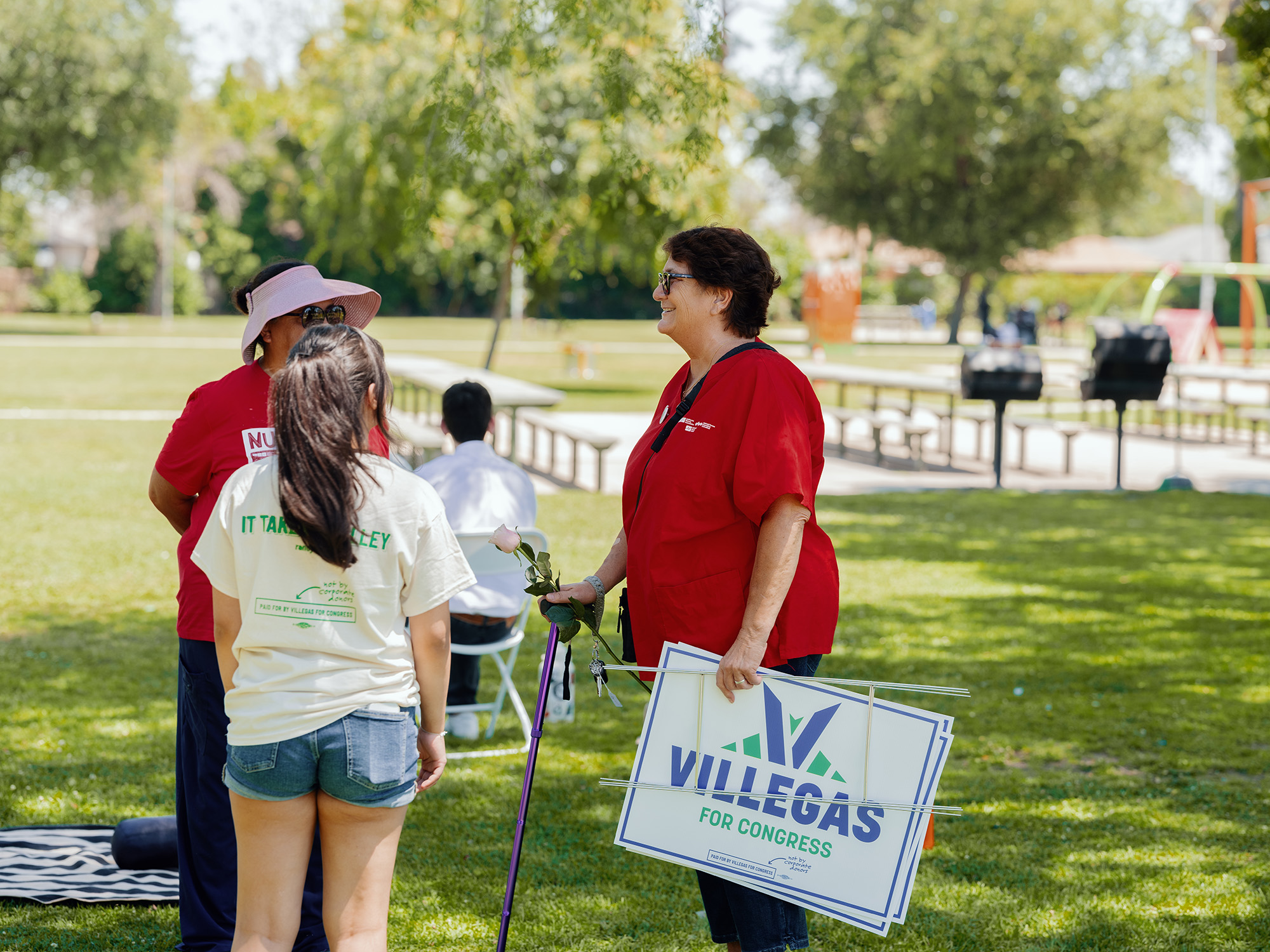 Three women stand in a grassy park talking. One holds a "Villegas for Congress" yard sign.