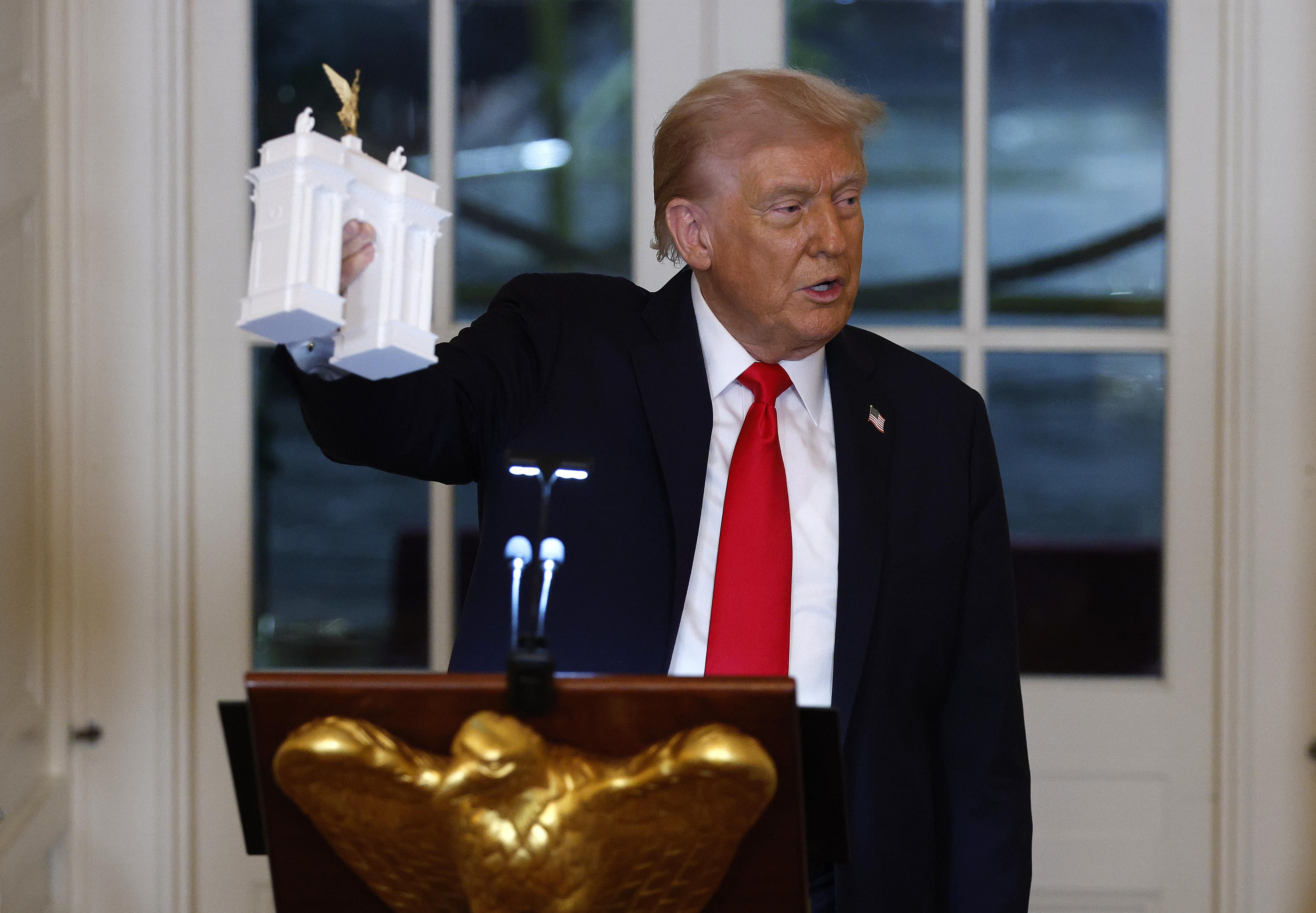Donald Trump, wearing a suit and tie, holds a white plastic model of an arch as he speaks from a podium decorated with a golden eagle.
