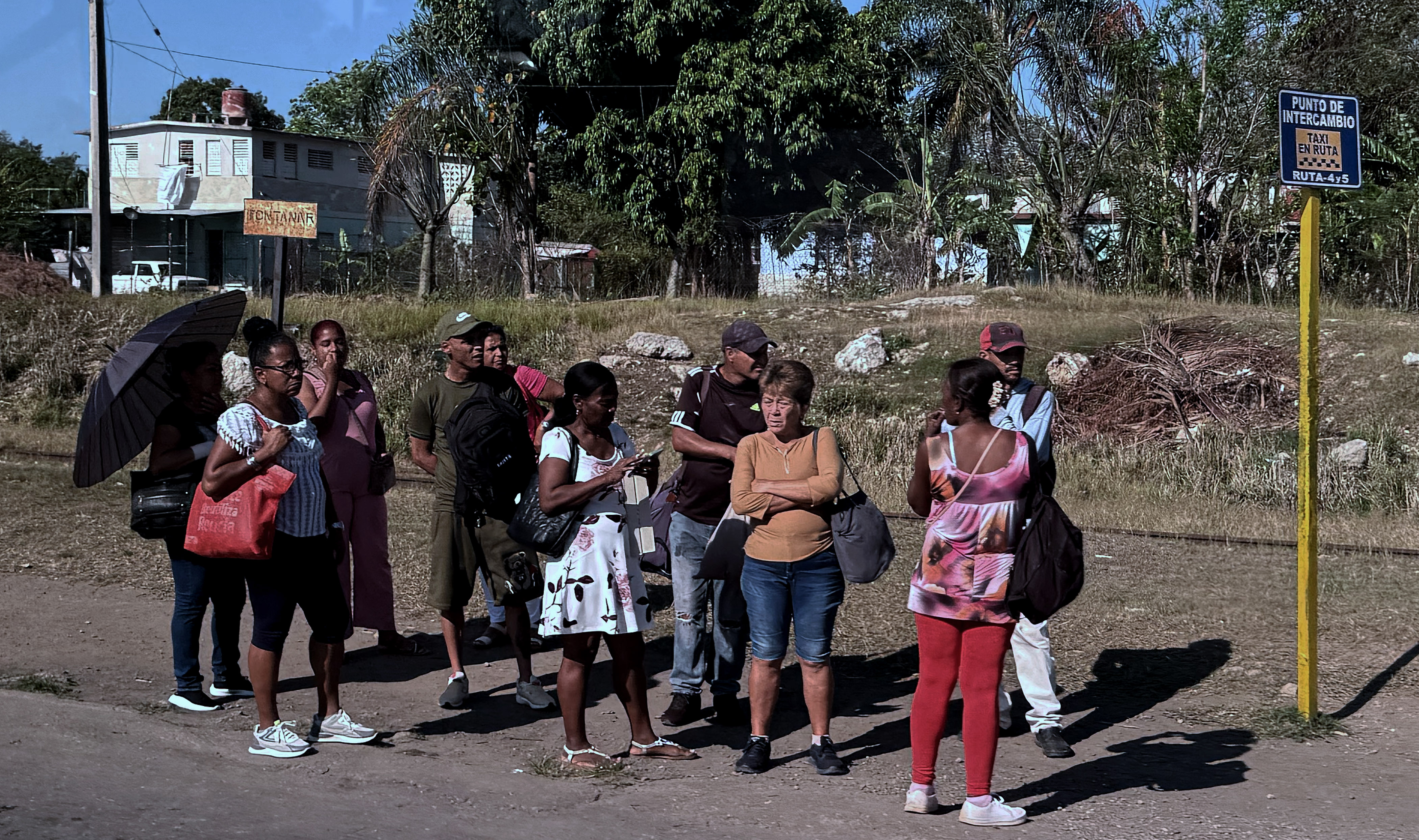 A line of people wait on a dirt road for a bus in the sun in Cuba.