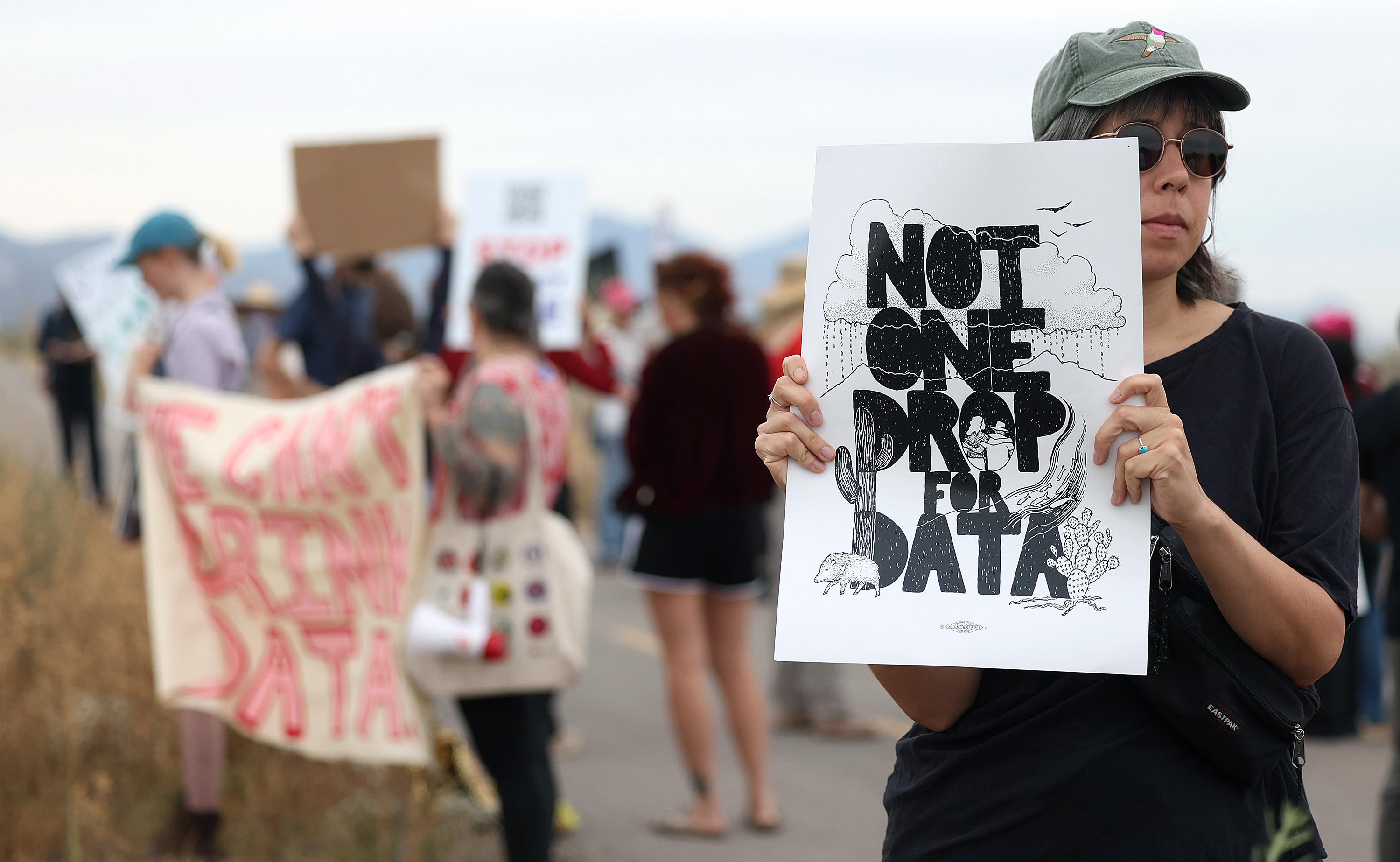 A protester wearing sunglasses holds a sign reading “Not one drop for data” during a roadside demonstration, with other protesters and signs blurred in the background.