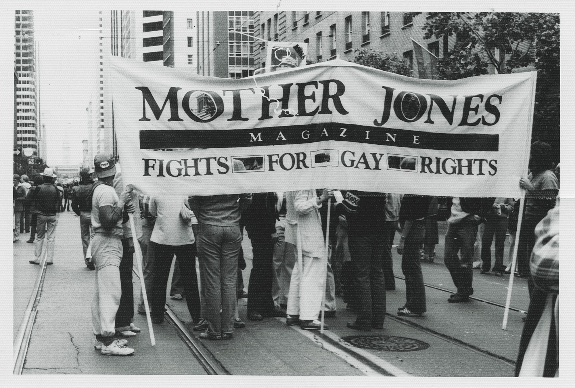 People stand in a street holding a banner that reads, "Mother Jones Magazine Fights For Gay Rights." The people's faces are obscured behind the banner.
