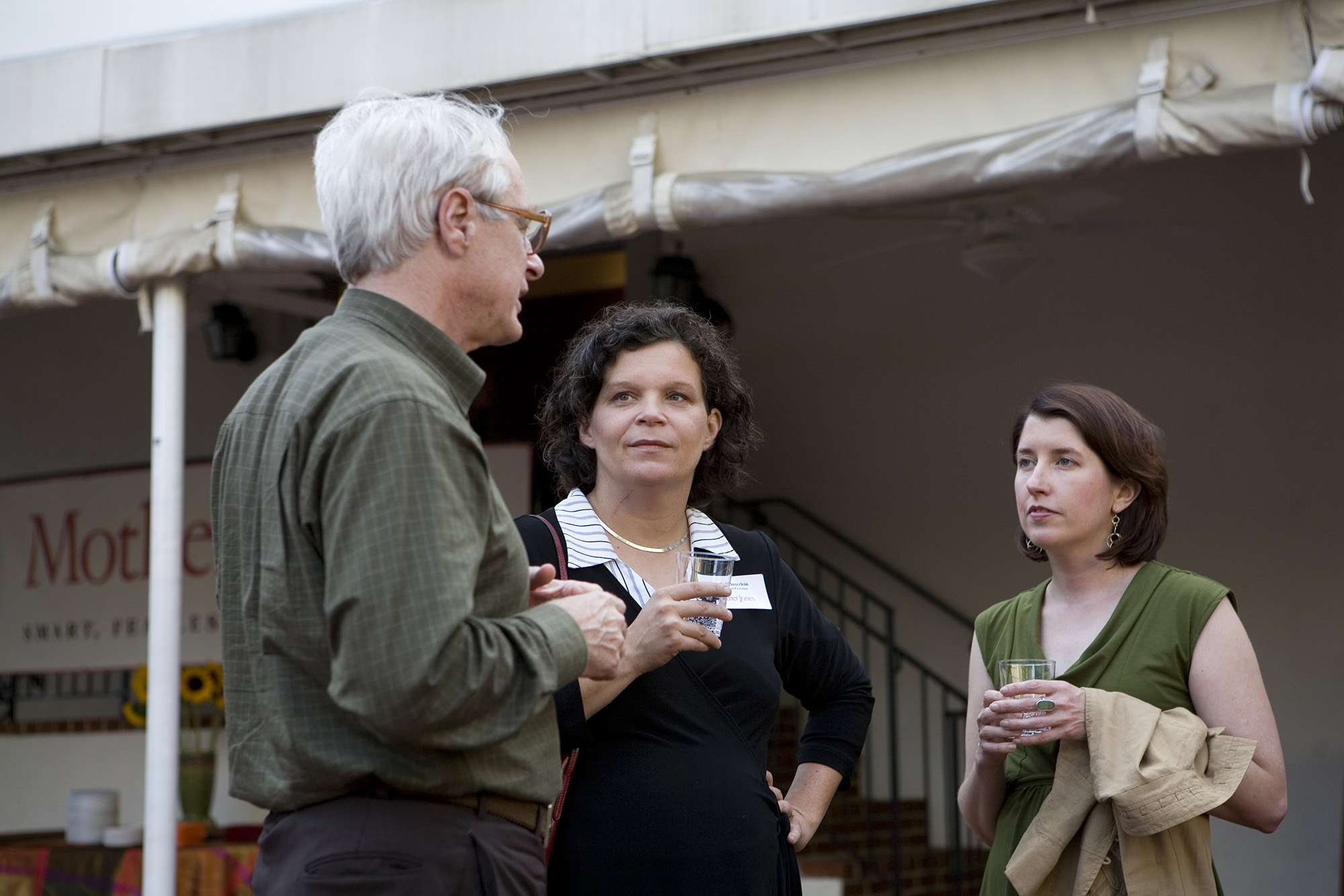 Two women stand facing a man seen in profile, who appears to be speaking.