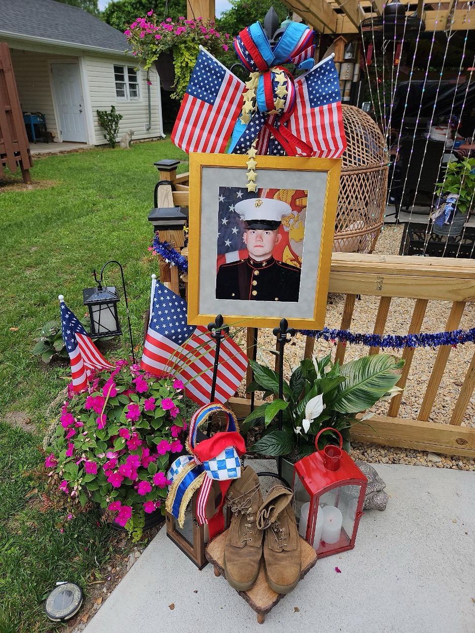 A photo of Drew in his Marine uniform with flowers, American flags, a pair of brown boots, and lanterns surrounding it.