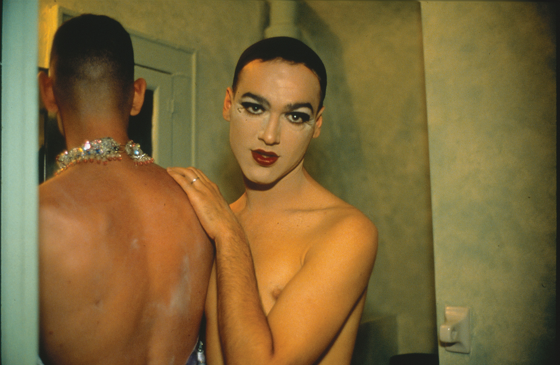 Two shirtless people, shown from the waist up, in the midst of changing into drag. One, seen from behind, wears a heavily jeweled necklace. One faces the camera, wearing a soft expression and heavy, glamorous makeup.