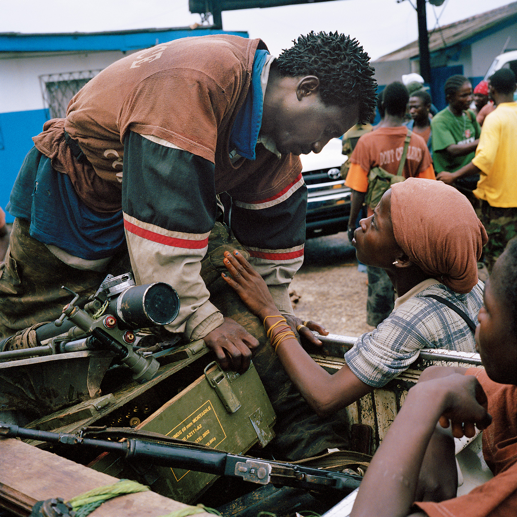 A young man and woman gaze lovingly at each other, surrounded by weapons and other people in the background.