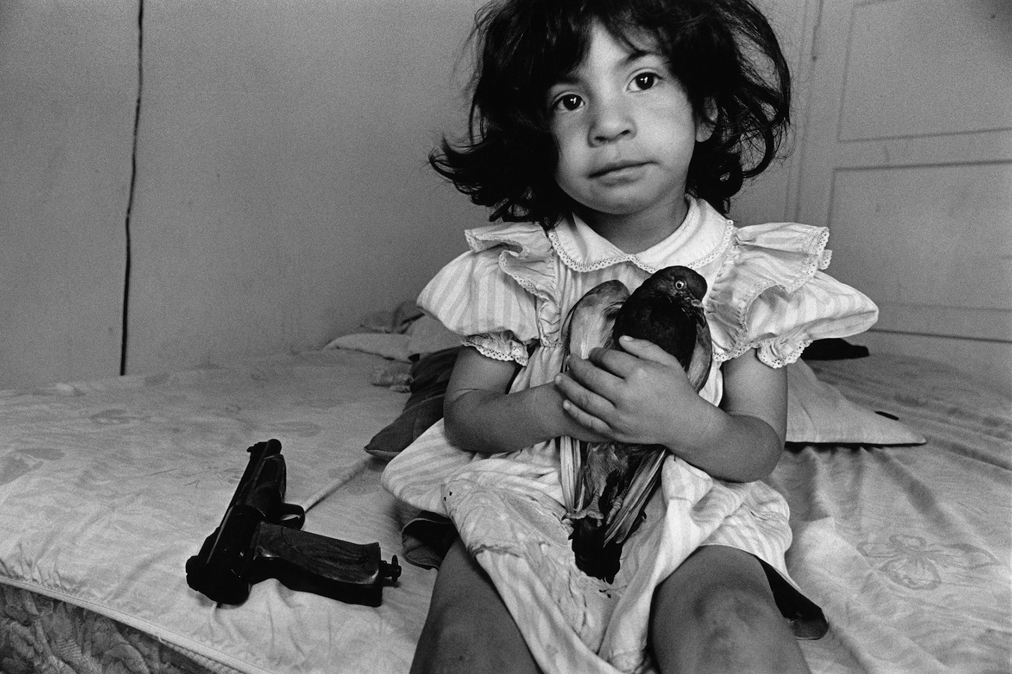 A black-and-white portrait of a young girl, seated on a mattress, clutching a bird. A handgun sits beside her on the bed.