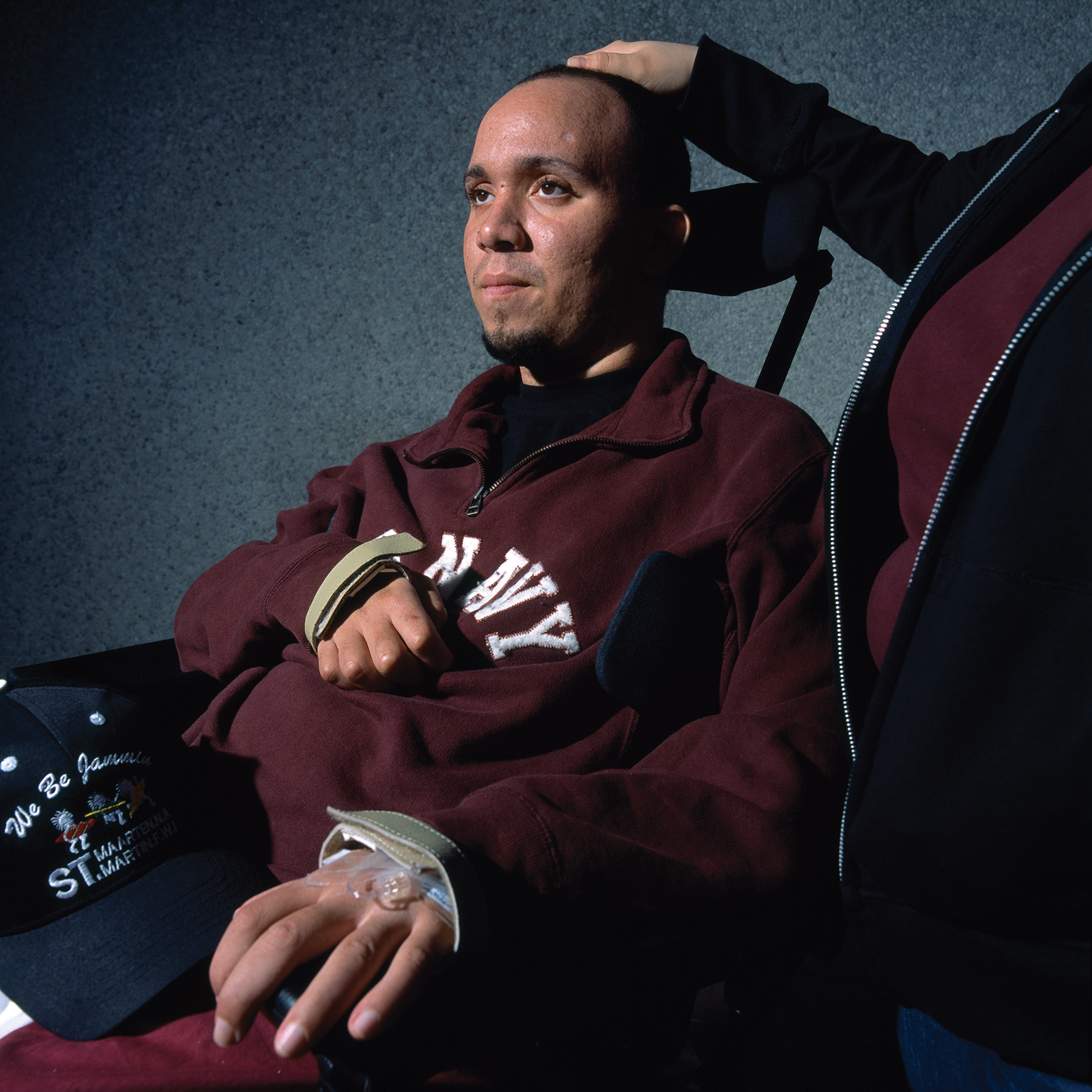 A close-up of a man with close-cropped dark hair in a wheelchair, who gazes off into the distance. An IV line is attached to his left hand.