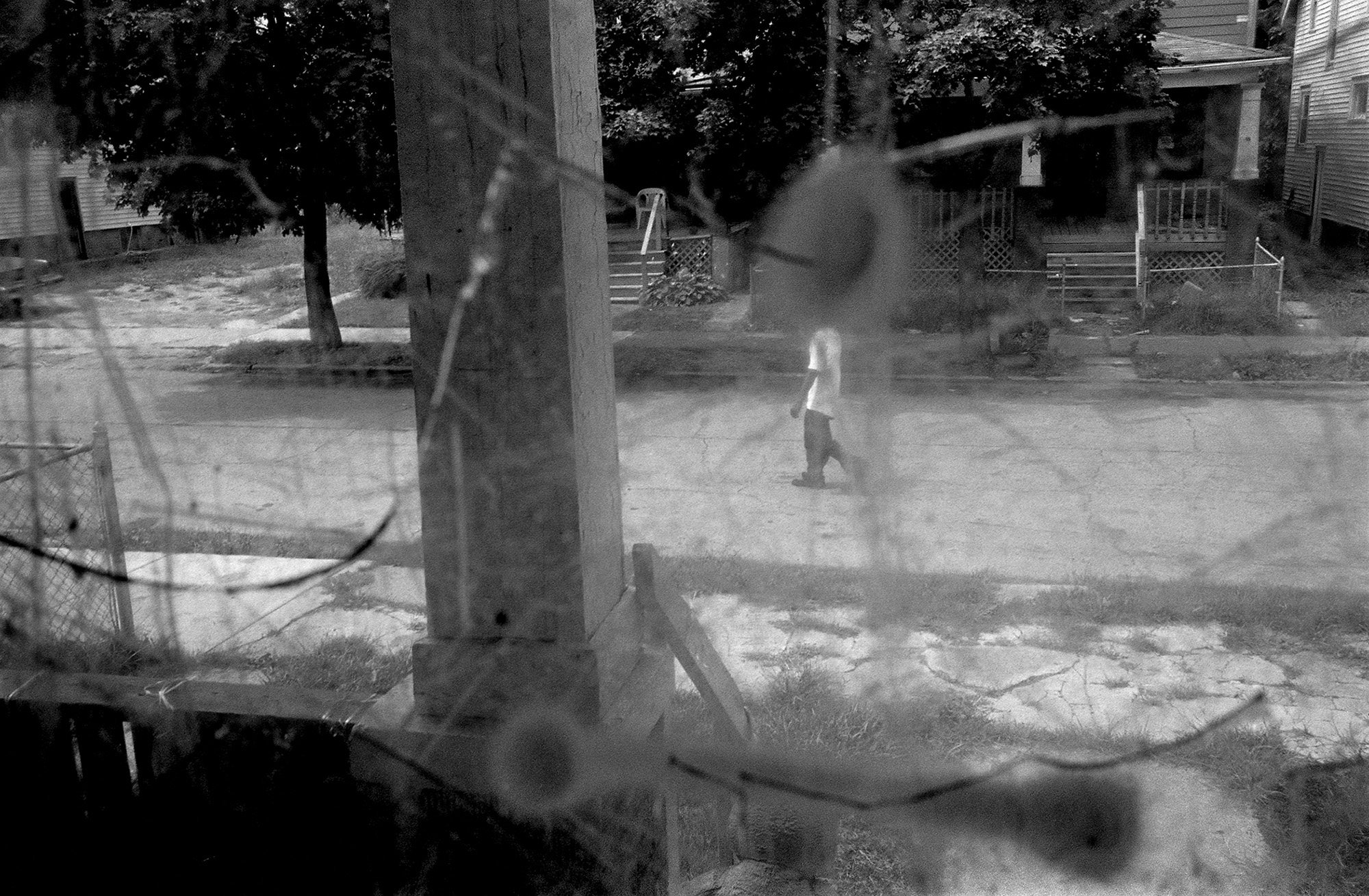 A black-and-white photo of a man walking down a street, as seen through a cracked window riddled with bullet holes.