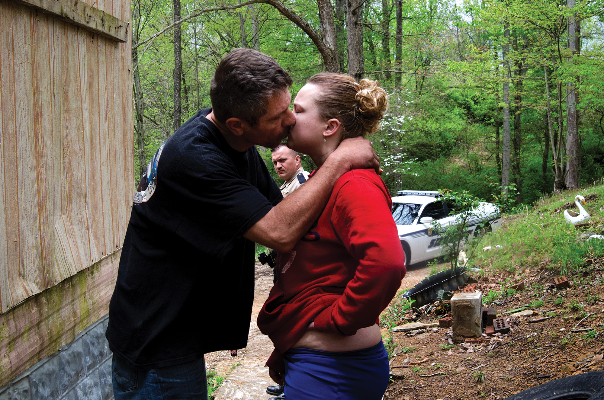 A couple kiss near a dilapidated structure in the woods. Behind the couple, a law enforcement officer and his squad car are visible.