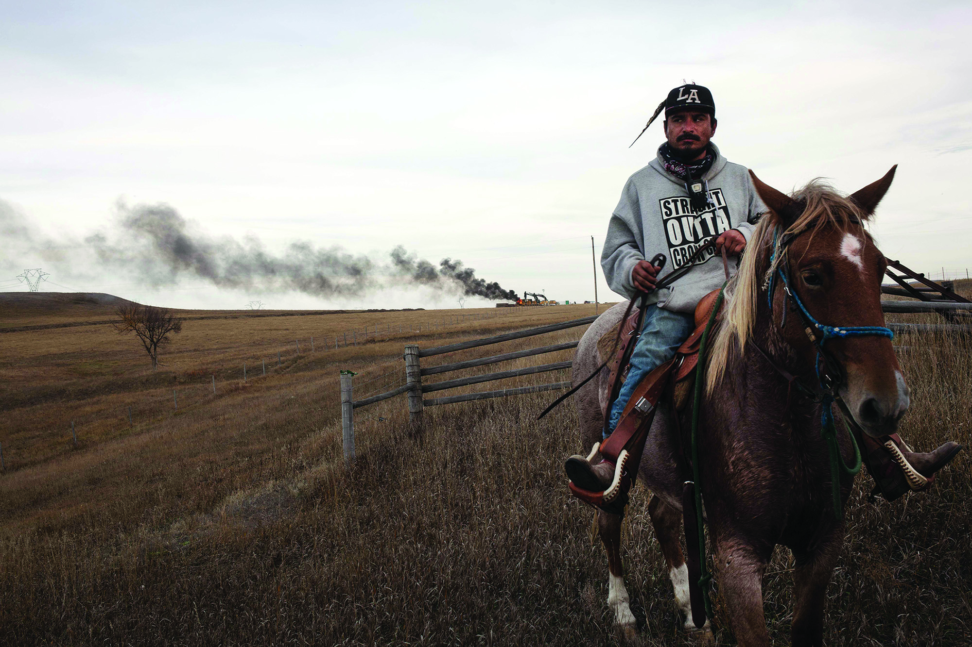 A young man wearing a gray hooded sweatshirt and dark baseball hat sits astride a horse in an open field. In the distance, black smoke billows out.