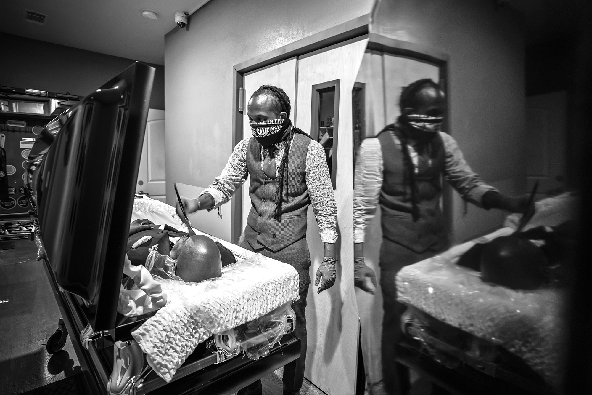 A black-and-white photo of a man, dressed in vest, tie, and long-sleeved shirt, attending to a body in an open casket. The man also has a large cloth tied across his face to cover his nose and mouth.