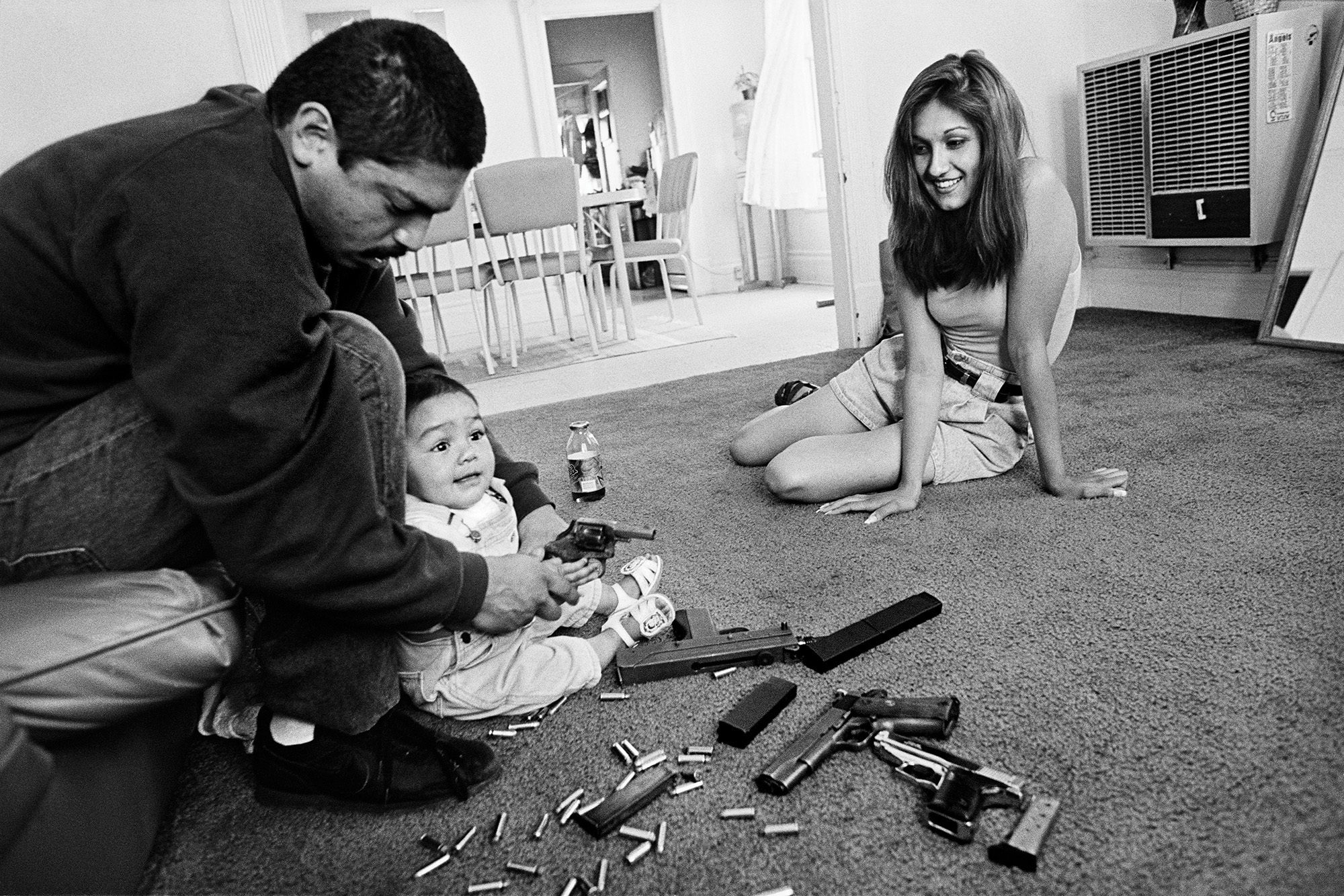 A black-and-white photo of a young family in their living room. A smiling woman sits on the carpeted floor, while a man sits on a low couch with a baby seated on the floor between his legs. The man helps the baby hold a small pistol, and guns, magazines, and bullets are strewn across the floor.