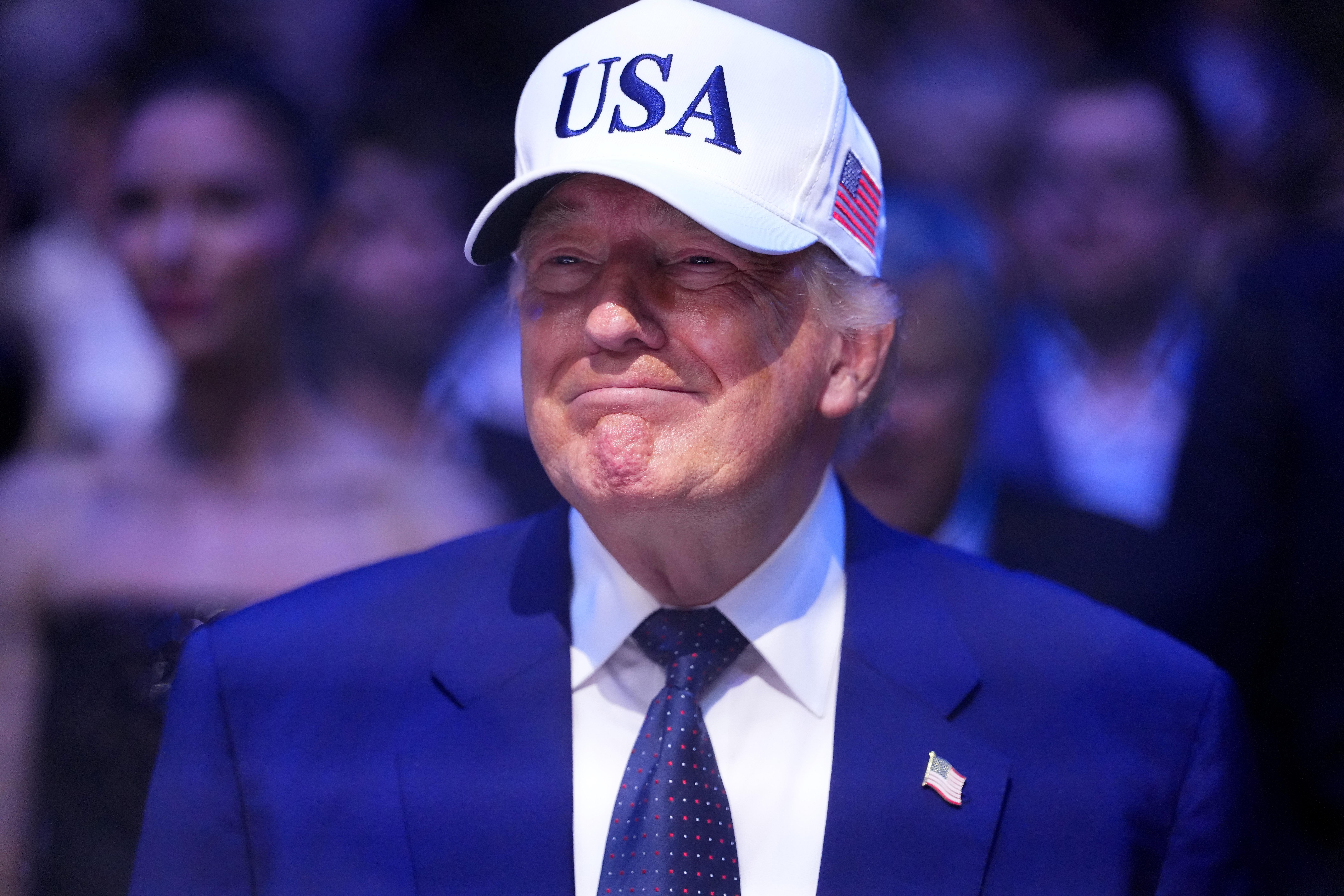 Donald Trump, wearing a suit and tie with a white “USA” ballcap, smiles.