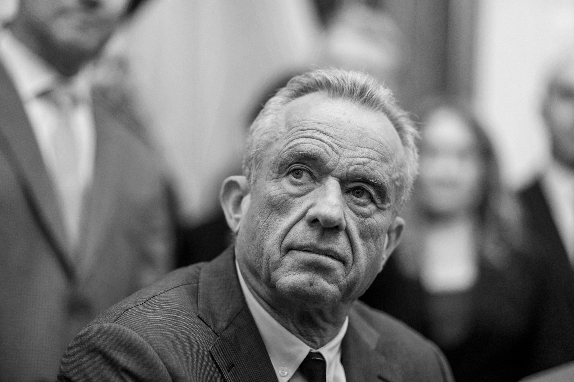 An elderly man with graying hair in suit and tie sits as people are gathered around him.