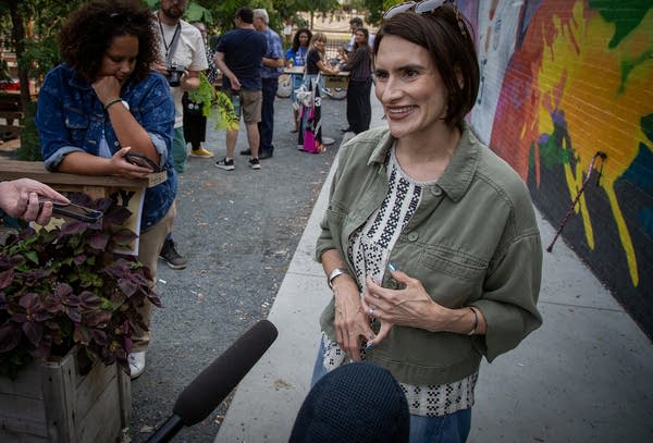 A woman smiles while talking to reporters in front of a mural on a bar patio.