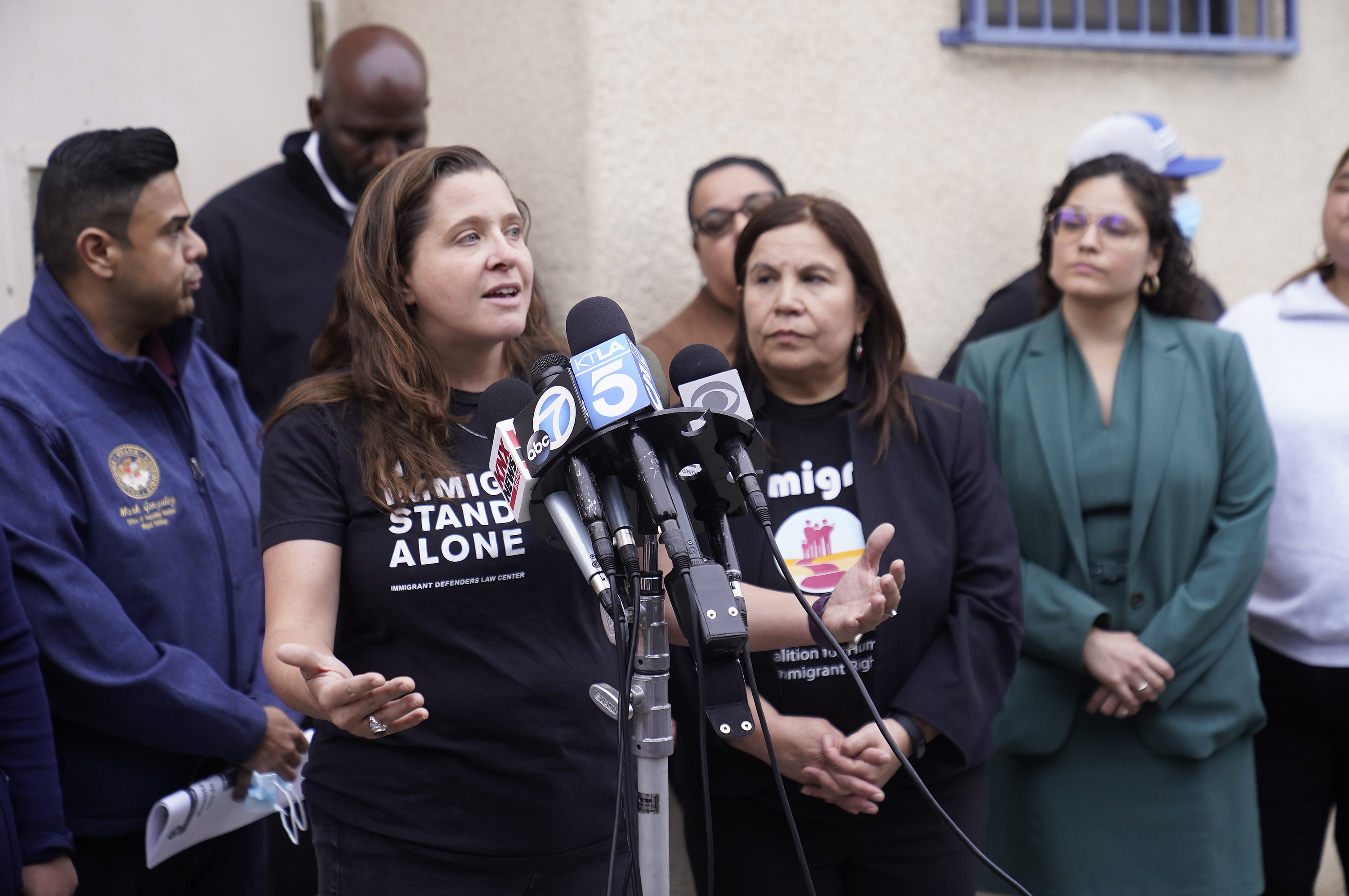 A woman wearing a T-shirt reading "NO IMMIGRANT STANDS ALONE" gestures while speaking into news microphones during an outdoor press conference.