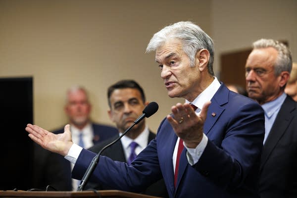 A man in a navy suit and red tie speaks at a podium in front of a group of government officials.