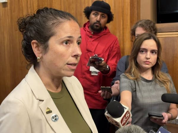 A woman speaks into several microphones held by reporters.