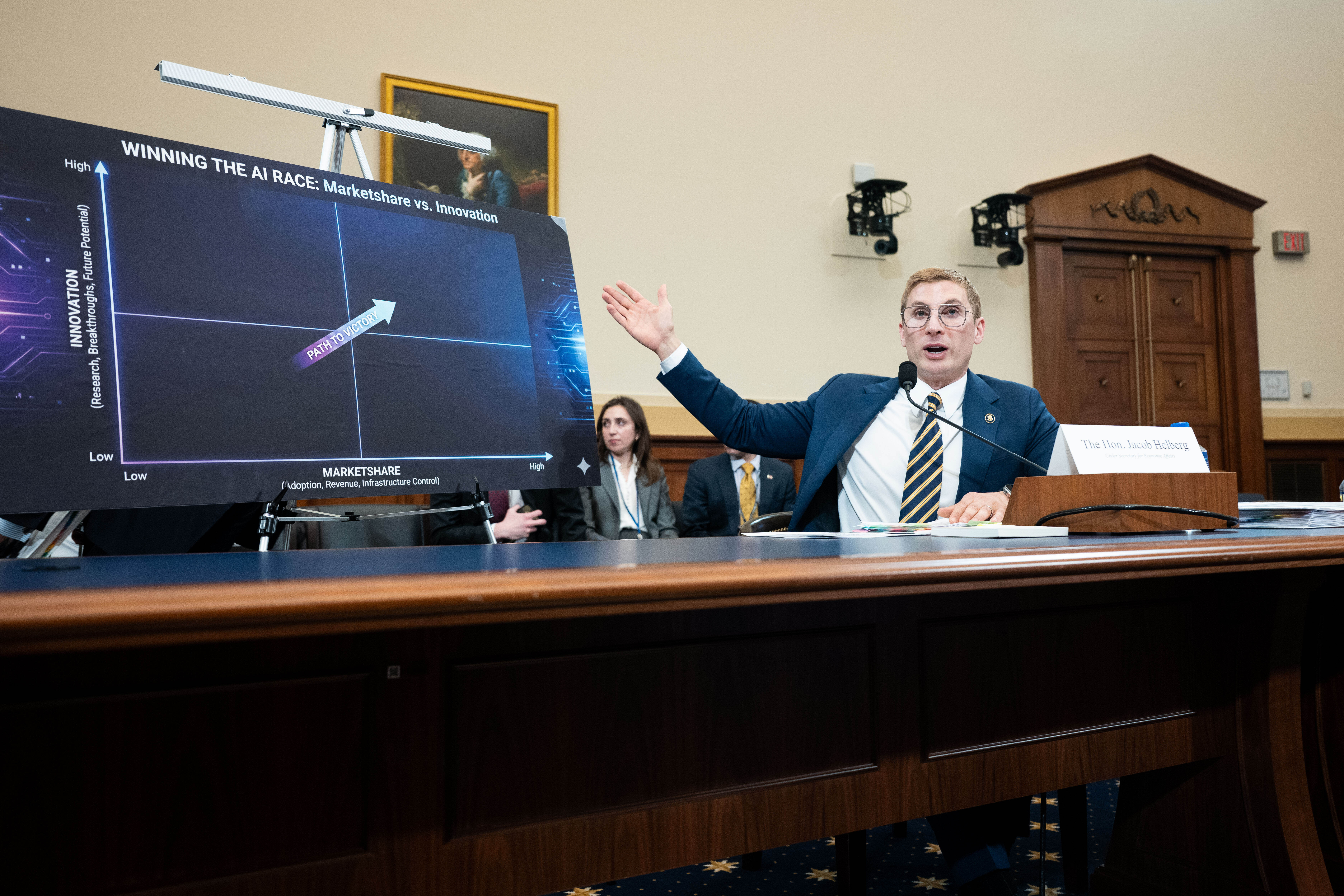 Jacob Helberg wearing a suit and glasses gestures to a graphic about AI economic growth. 