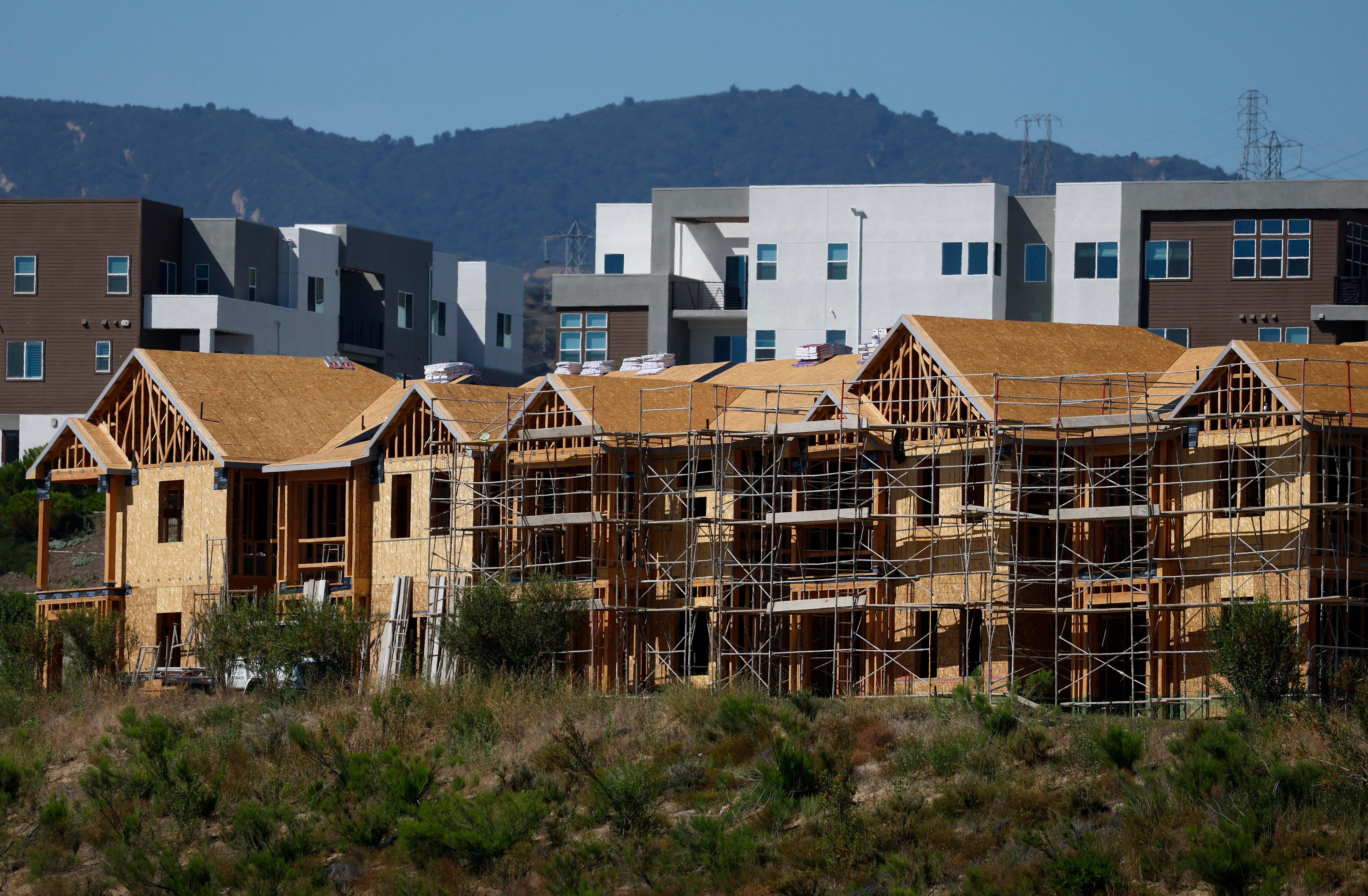 Rows of wood-framed homes under construction, surrounded by scaffolding, with newer apartment buildings and tree-covered hills in the background.