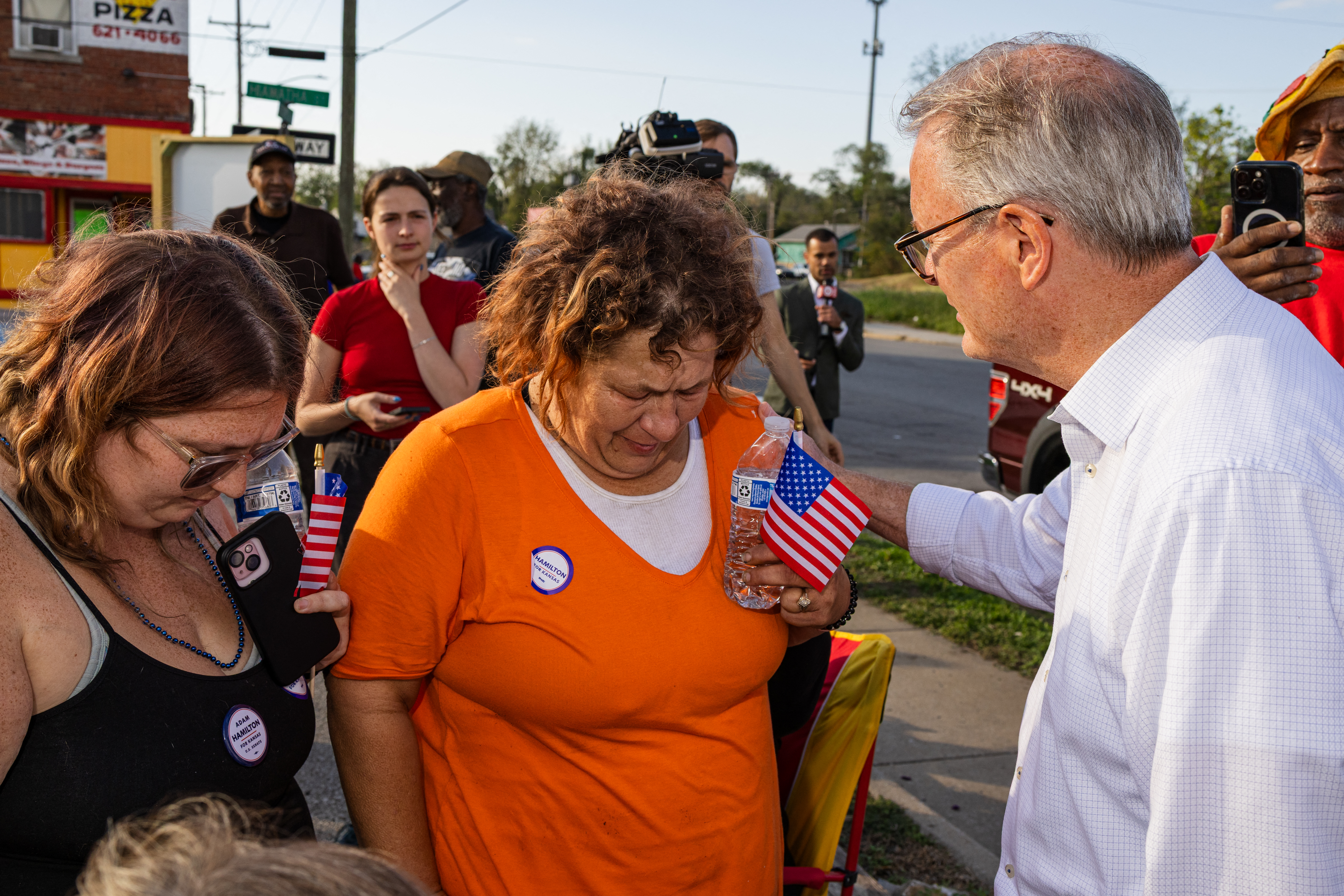 A man places his hand on the shoulder of a tearful woman holding a small US flag on a street. She wears a campaign button reading "HAMILTON FOR KANSAS 2024."
