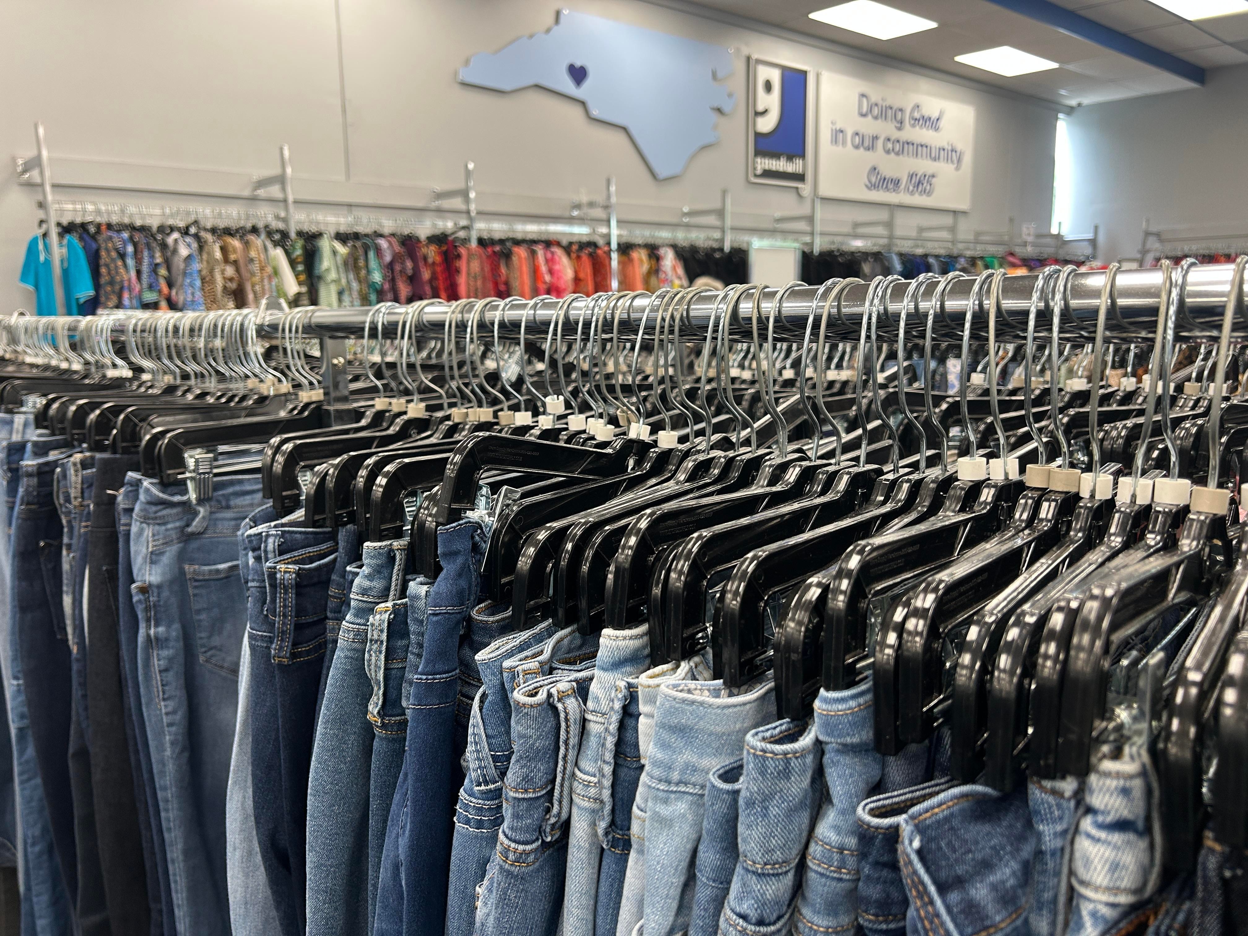A clothing rack filled with jeans in a Goodwill store; colorful shirts are visible on another rack in the background.