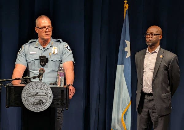 A uniformed police officer speaks at a podium in front of a dark blue curtain while a man in a suit stands next to him.