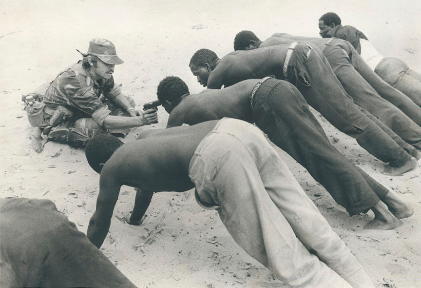 A white Rhodesian soldier points a gun at African villagers who are in a plank position during an interrogation.