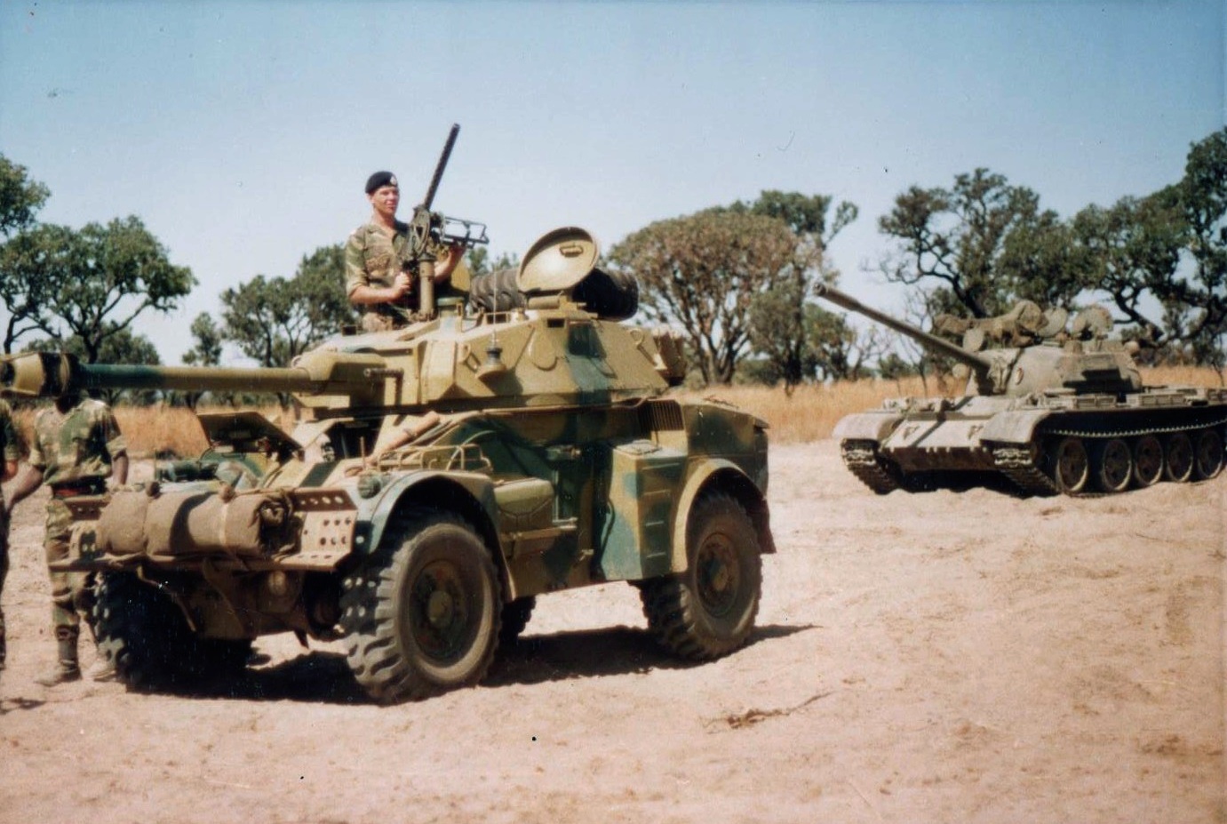 A Rhodesian Corps solider holds a gun and is visible at the top of an armored tank in desert scrubland.