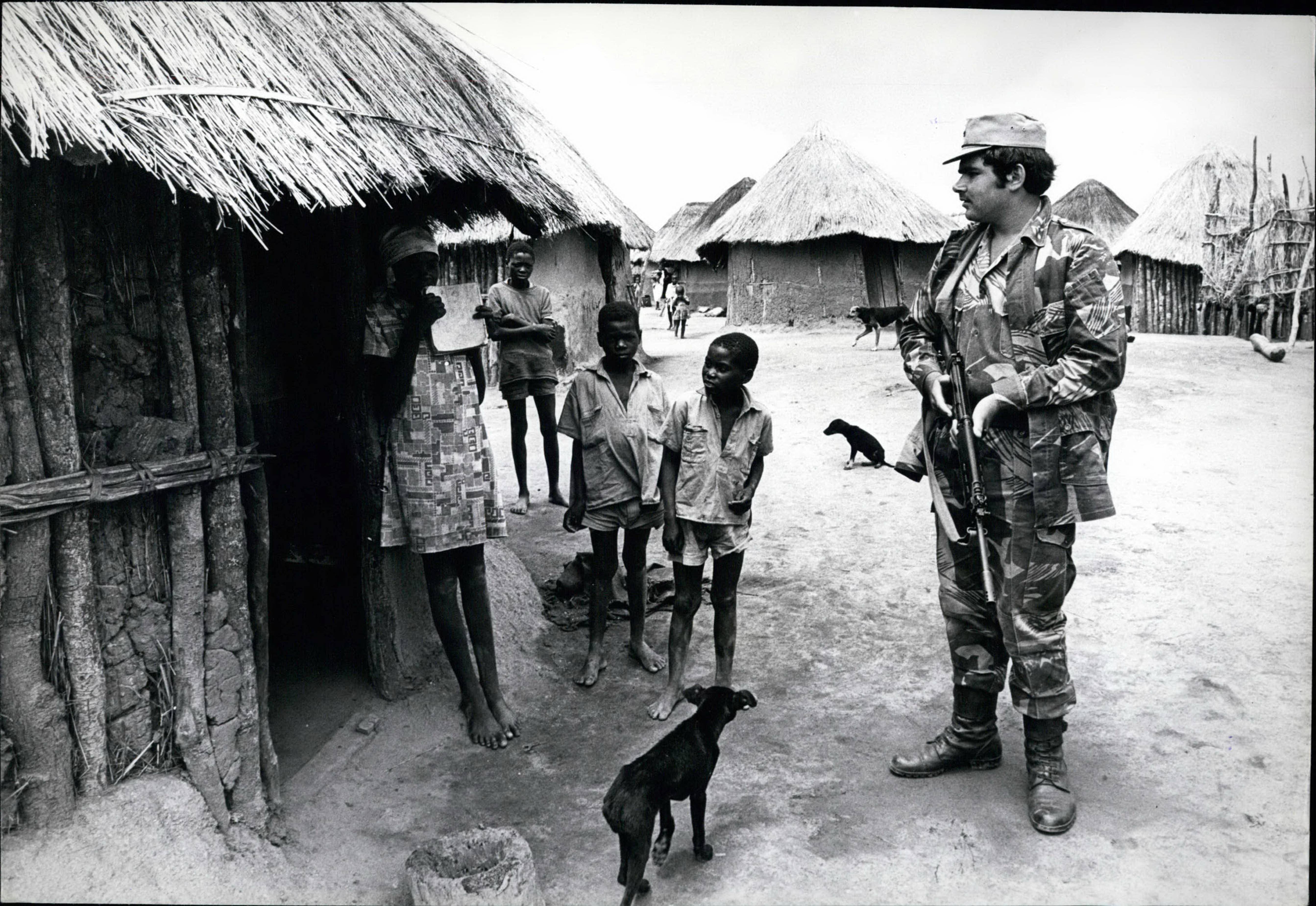A white soldier interrogates African villagers outside of their dwellings. 