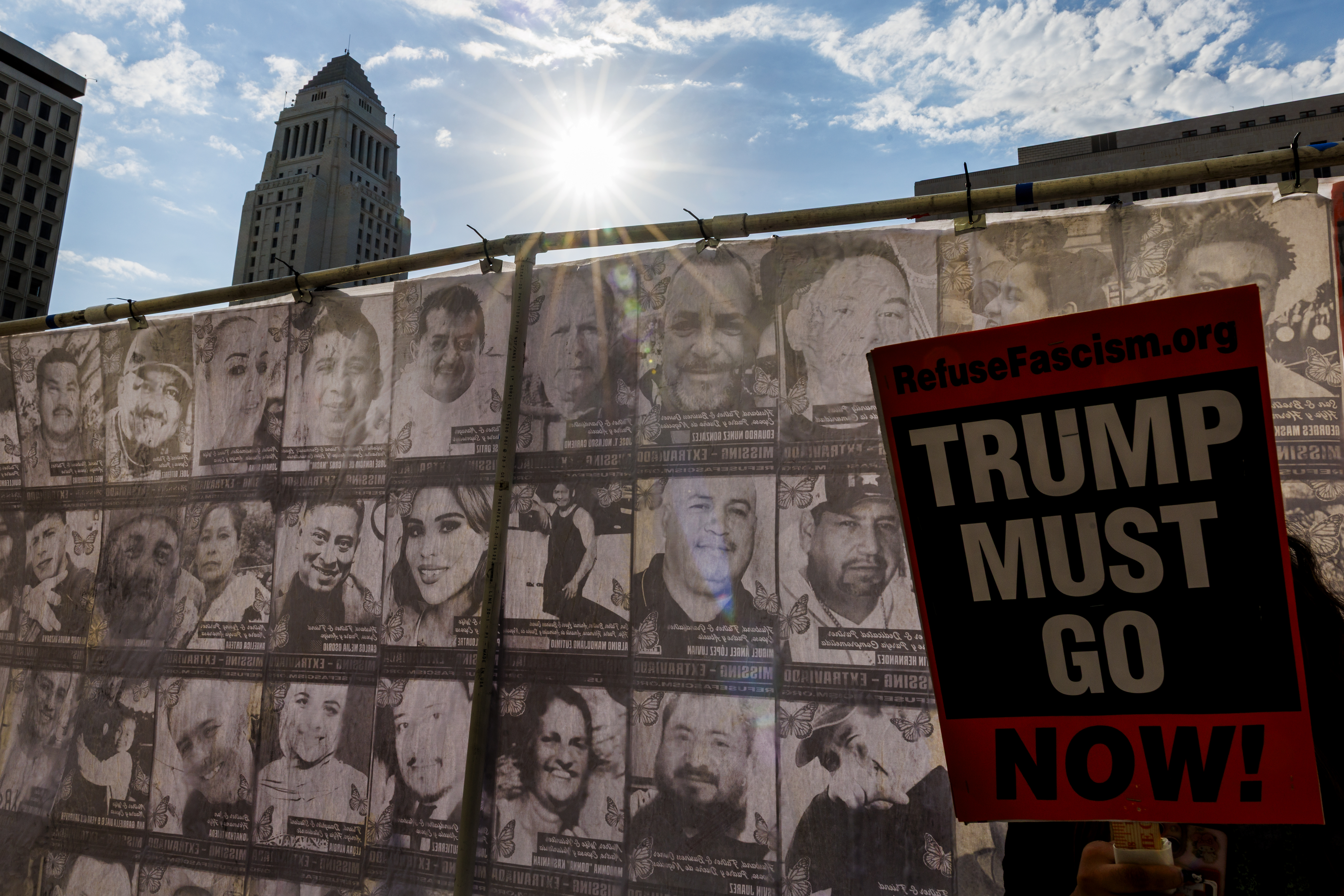 A photograph of a banner that has dozens of faces in black and white. Behind the banner stands the top of Los Angeles City Hall beside a shimmering sun. In front of the banner is a person eclipsed by the sign they are holding that reads "TRUMP MUST GO NOW!"