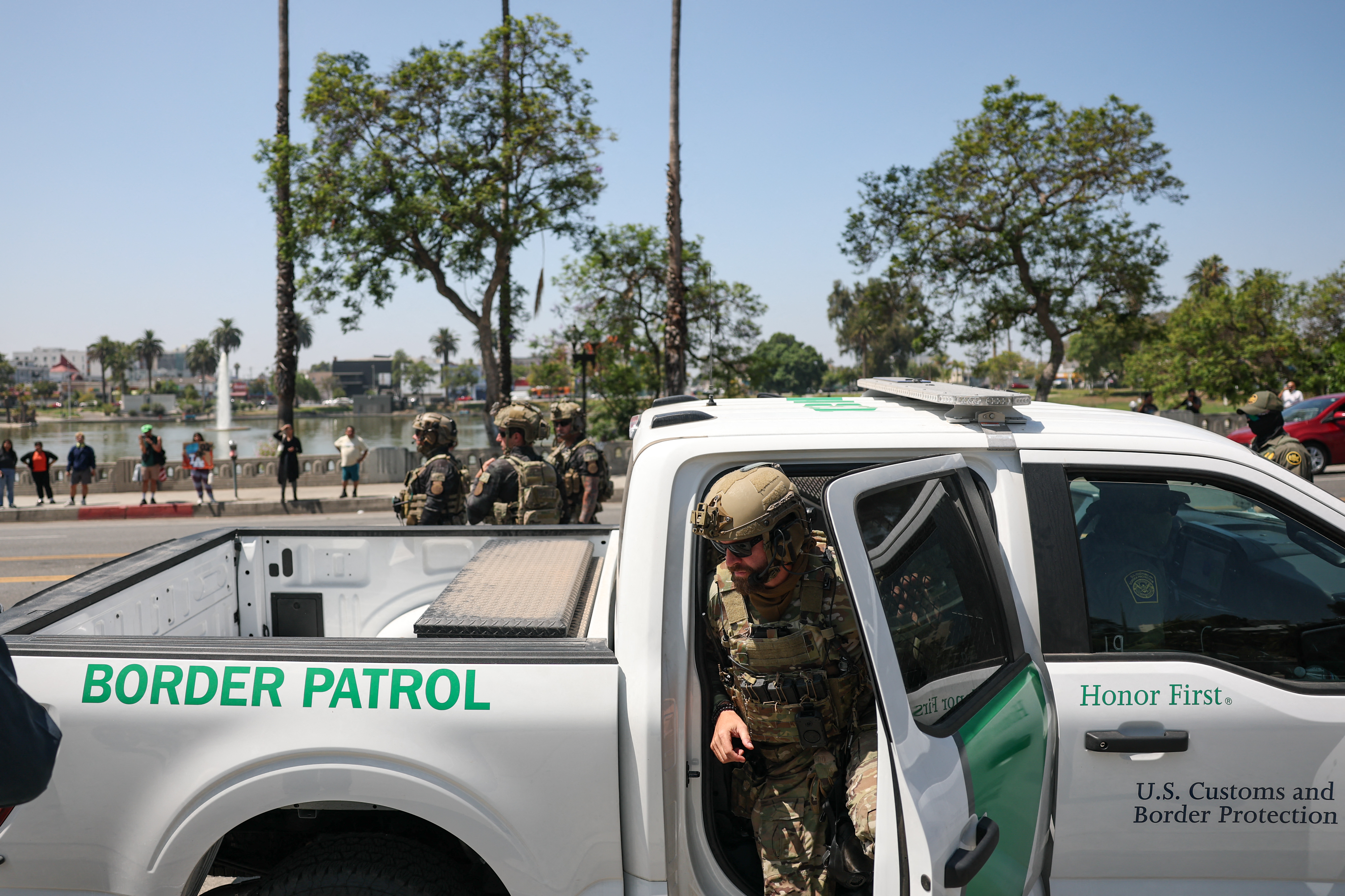 A photograph of an agent in military tactical gear getting out of a white pickup truck as other agents stand nearby in front of a park with trees and lake. The side of the white truck reads in part "BORDER PATROL" and "Honor First."
