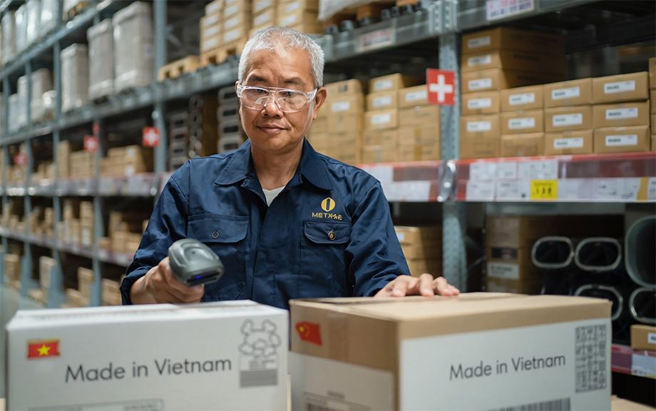 AI generated image of an Asian man in a warehouse with several shelves of cardboard boxes behind him as he scans two boxes in front of him getting ready to ship. Boxes say made in Vietnam.