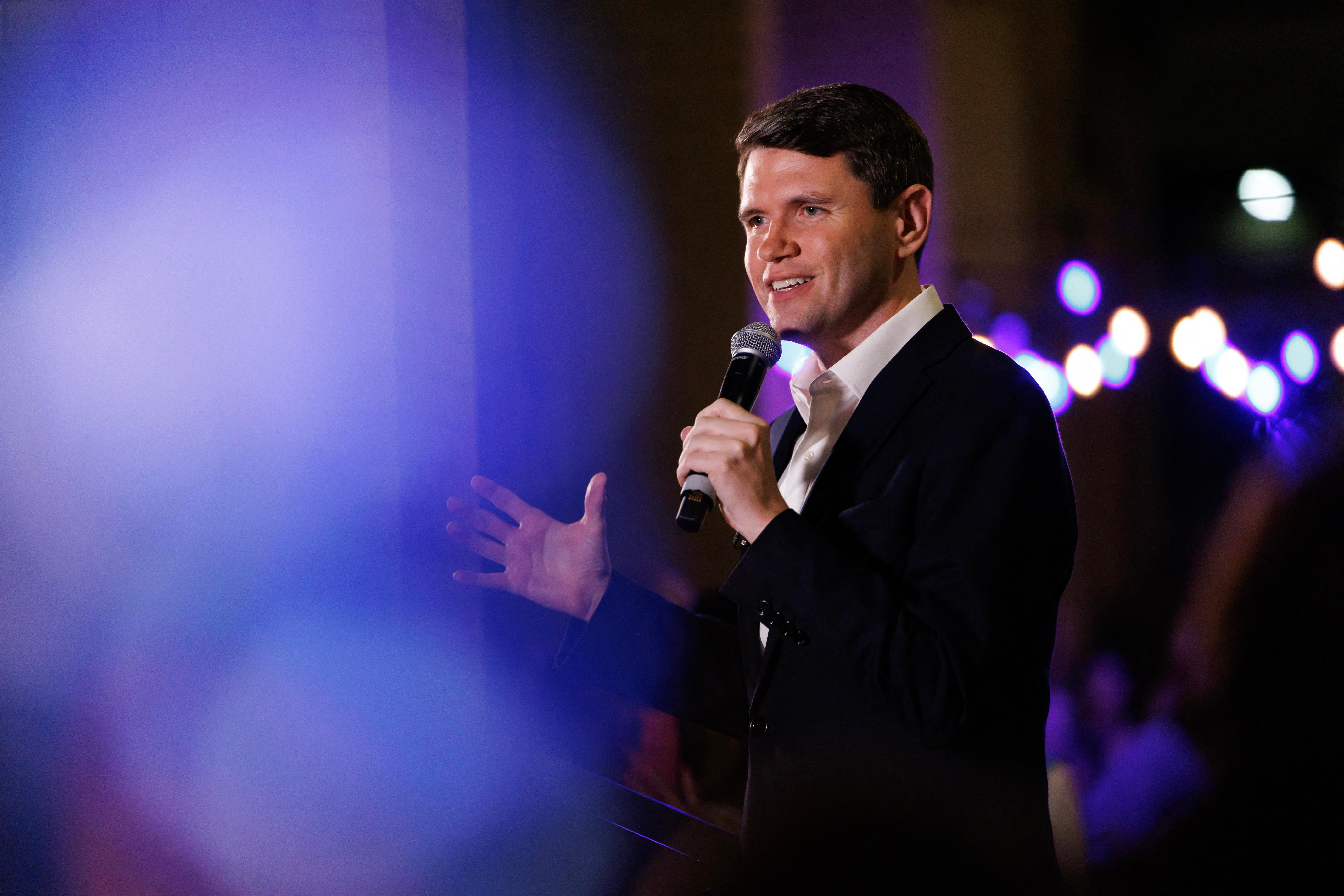 James Talarico gestures with one hand while speaking into a microphone; the left half of the frame is filled with blue lens flare.